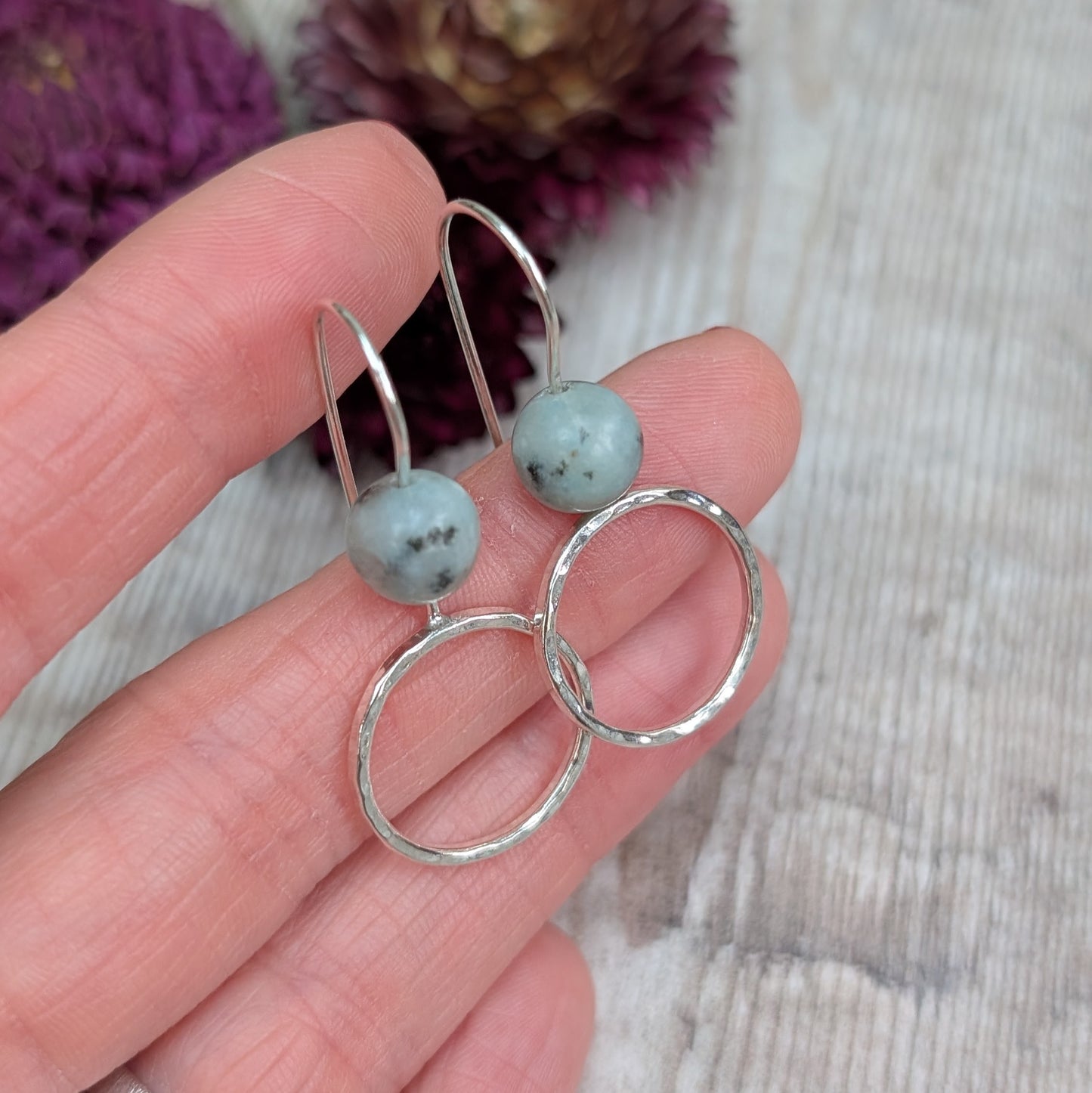 Hand holding a pair of Silver Hammered Circle Earrings with pale green Kiwi Jasper stones and hammered silver circles, with dried flowers in the background.