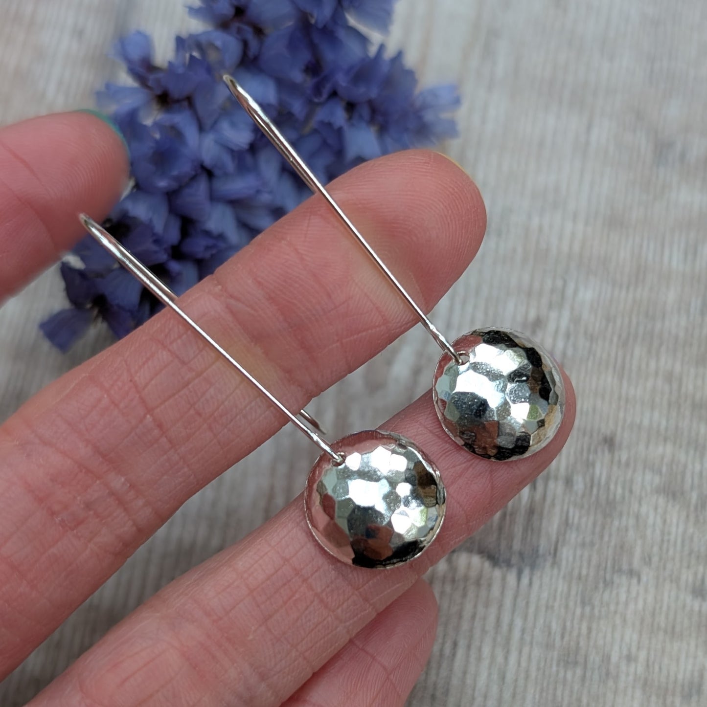Close-up of hammered silver long disc earrings held between fingers, showing the textured surface.