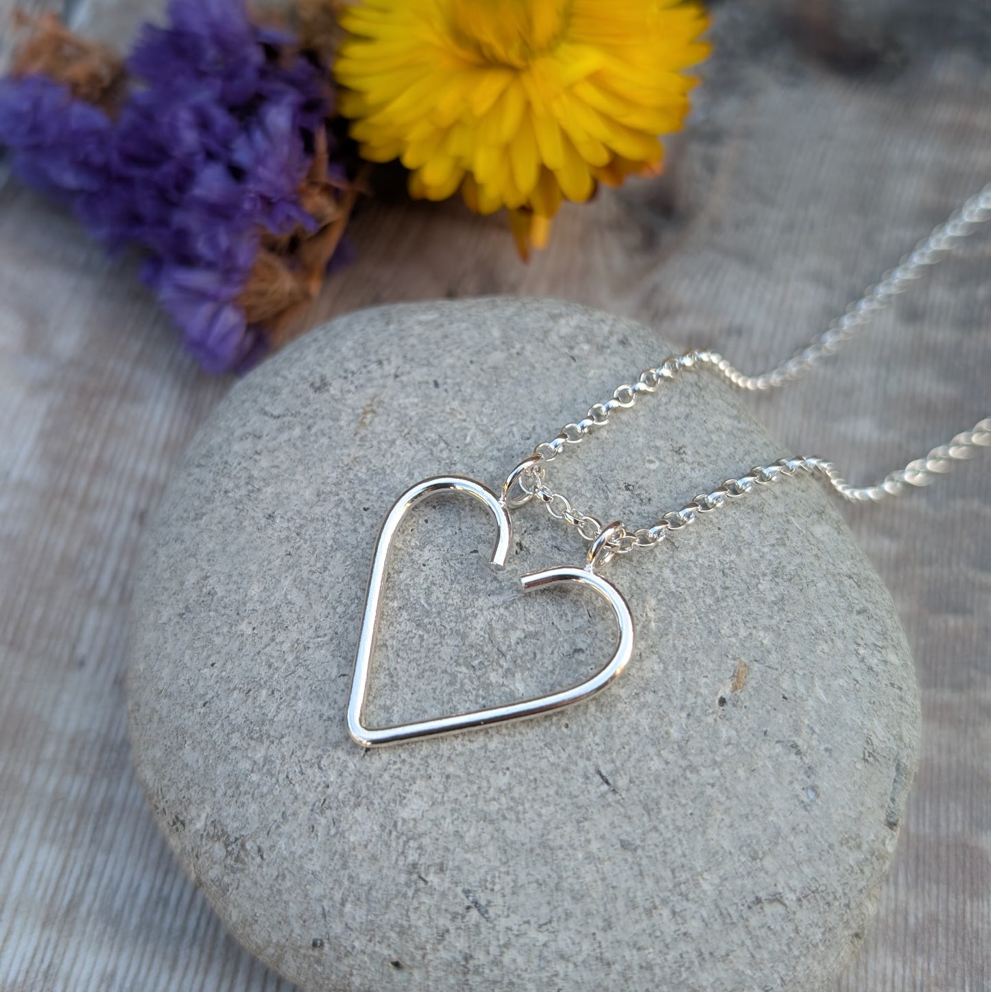 Silver heart ring keeper necklace displayed on a grey stone, with bright yellow and purple flowers behind it.