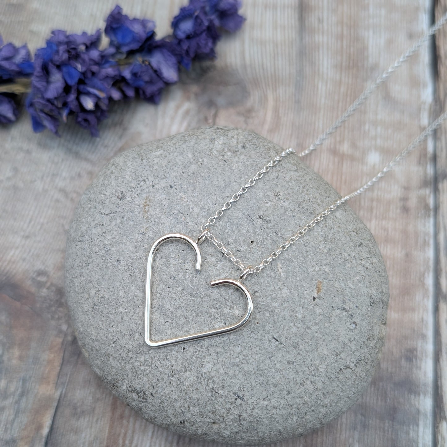 Silver heart ring keeper necklace placed on a grey stone with purple flowers in the background.