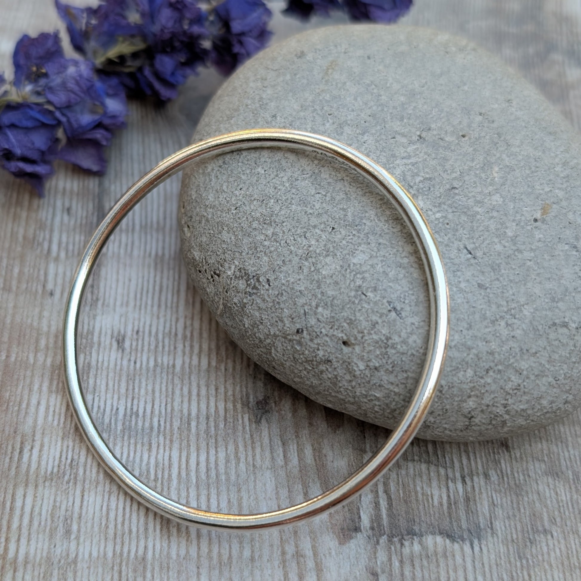 A silver round bangle resting at an angle on a grey stone, with a rustic wooden background and a few purple flowers visible.