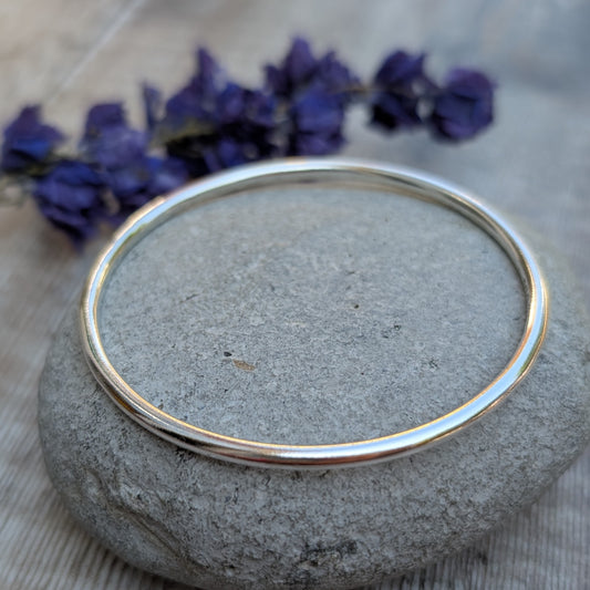A shiny silver round bangle displayed flat on a smooth stone, placed on a wooden surface with purple flowers nearby.