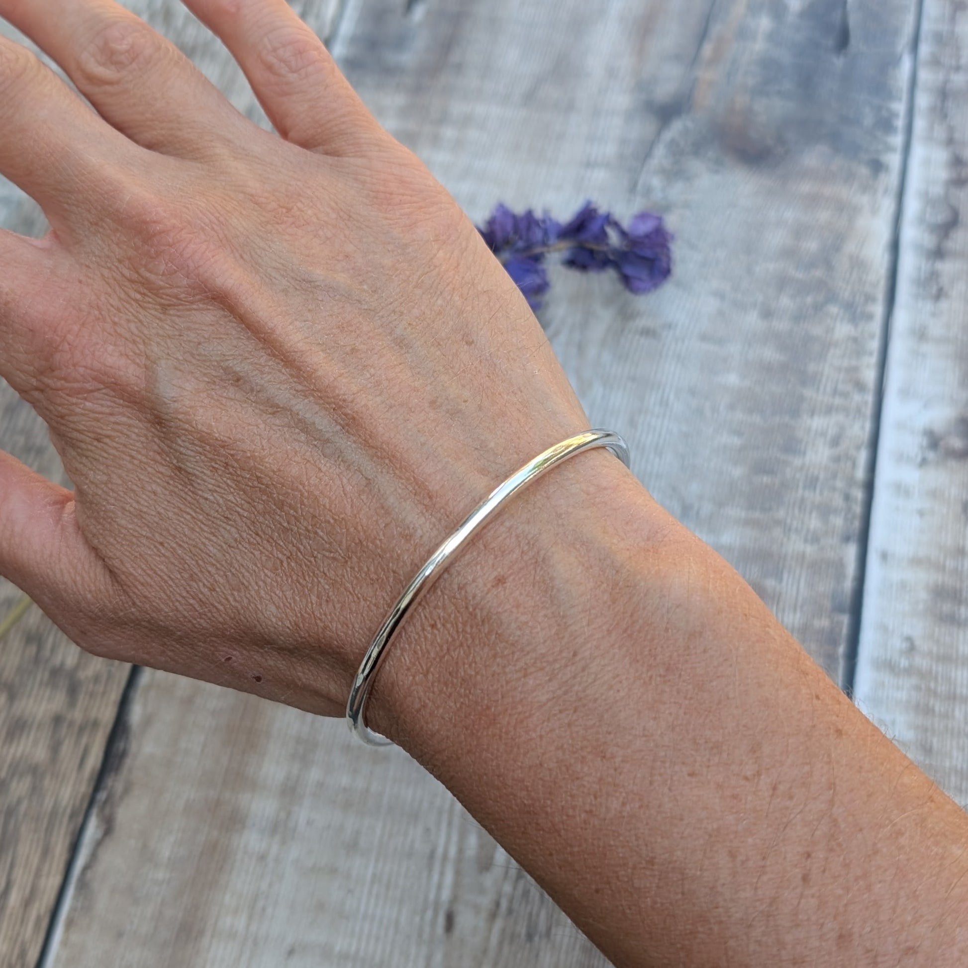 A silver round bangle worn on a wrist, with the bangle catching the light and purple flowers in the background.