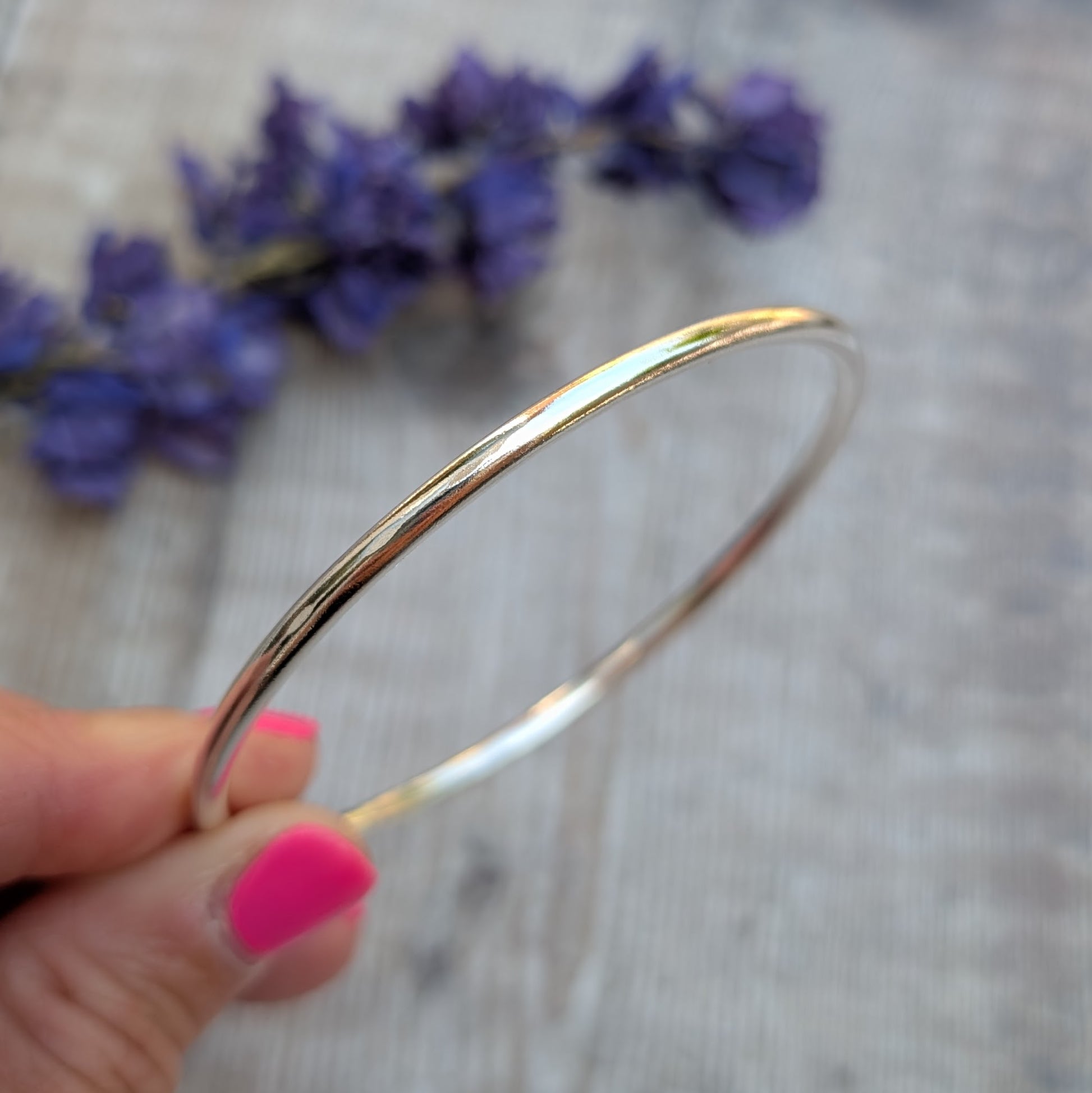 A close-up of a hand holding a simple, polished silver round bangle in front of blurred purple flowers.