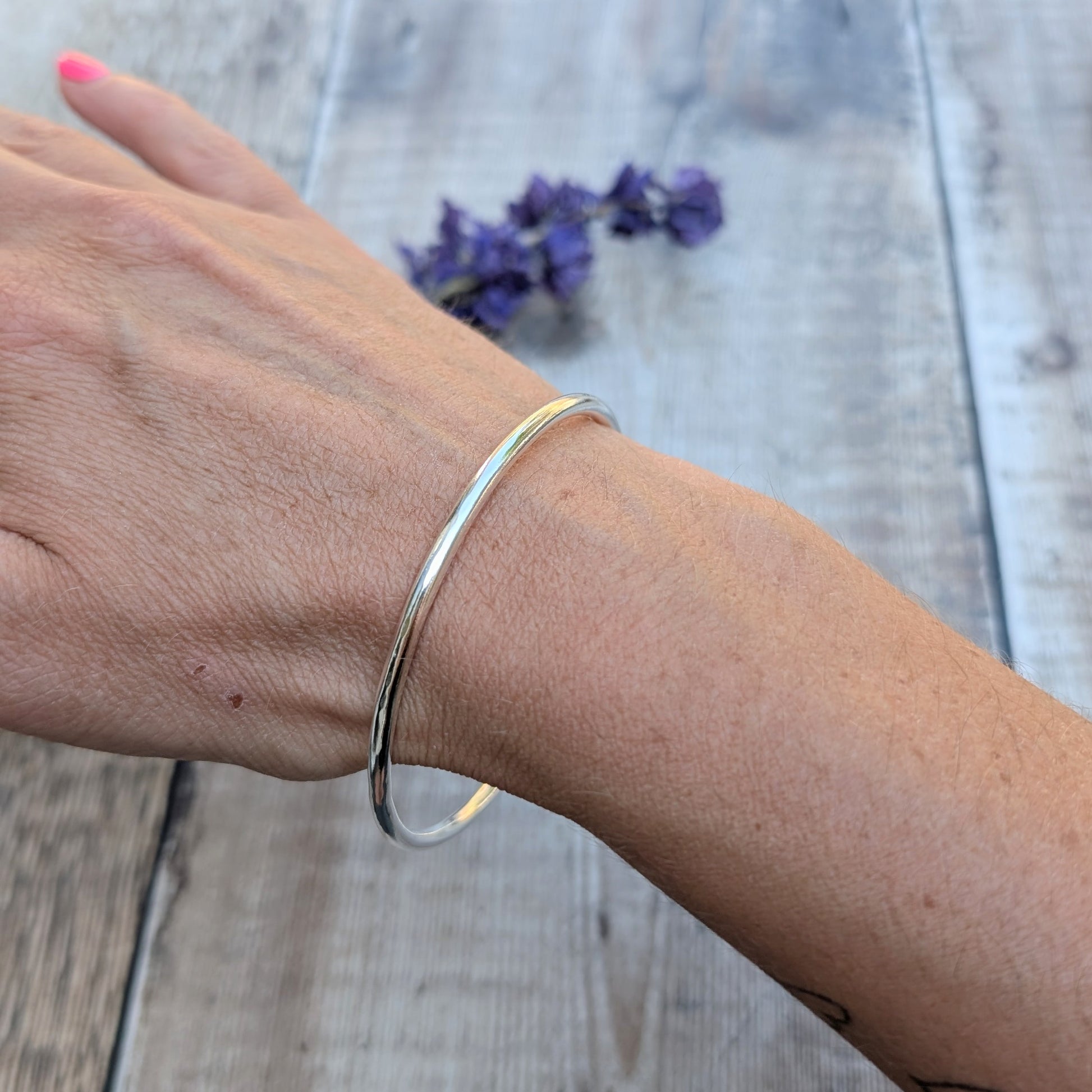 A silver round bangle worn on a wrist, with the bangle catching the light and purple flowers in the background.