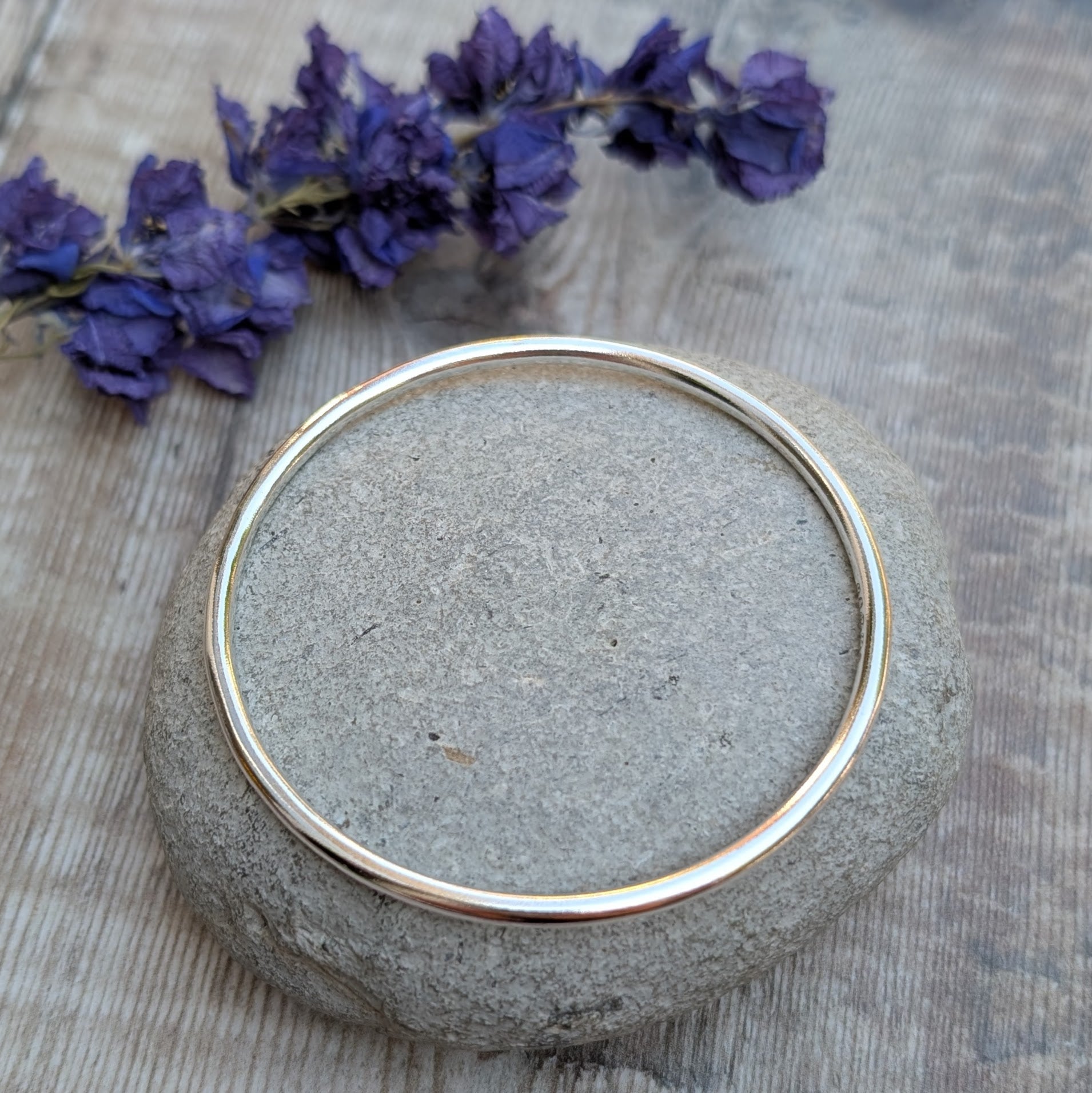 A shiny silver round bangle displayed flat on a smooth stone, placed on a wooden surface with purple flowers nearby.