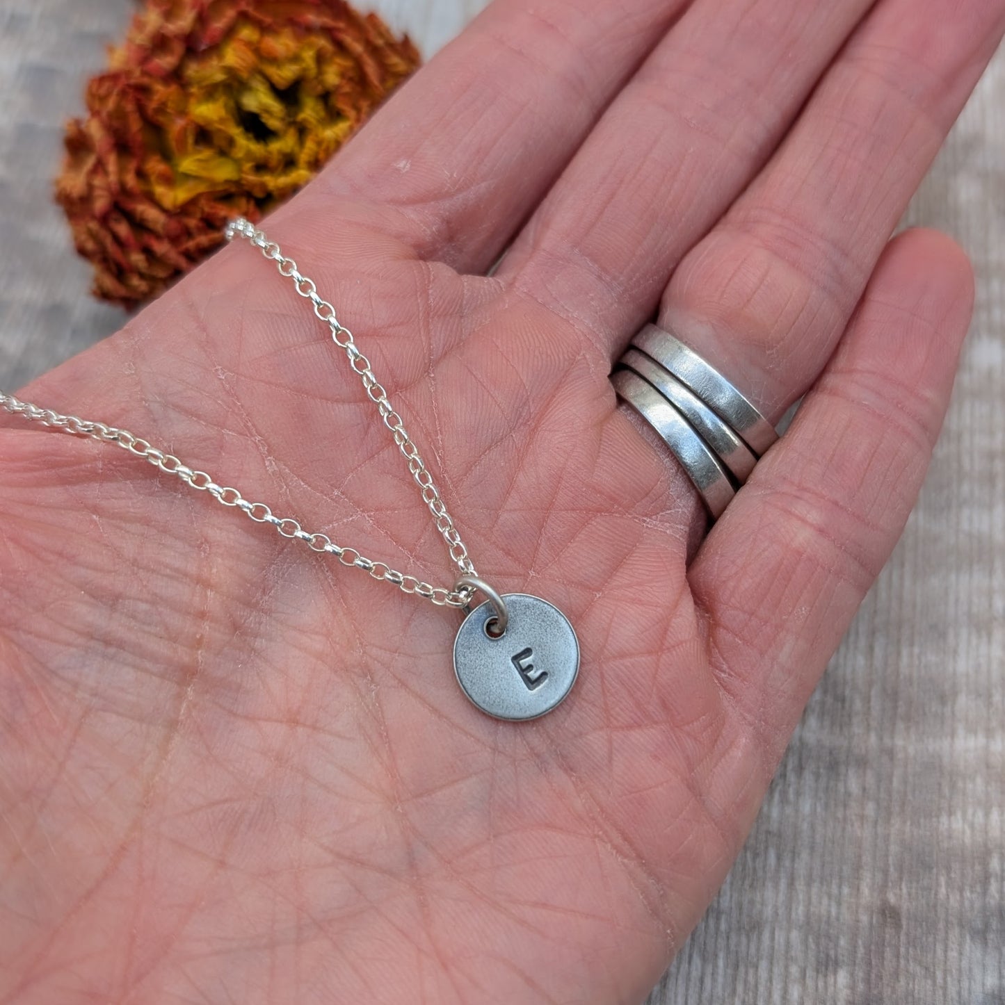 A hand holding a silver initial necklace with a single disc stamped with the letter "E."