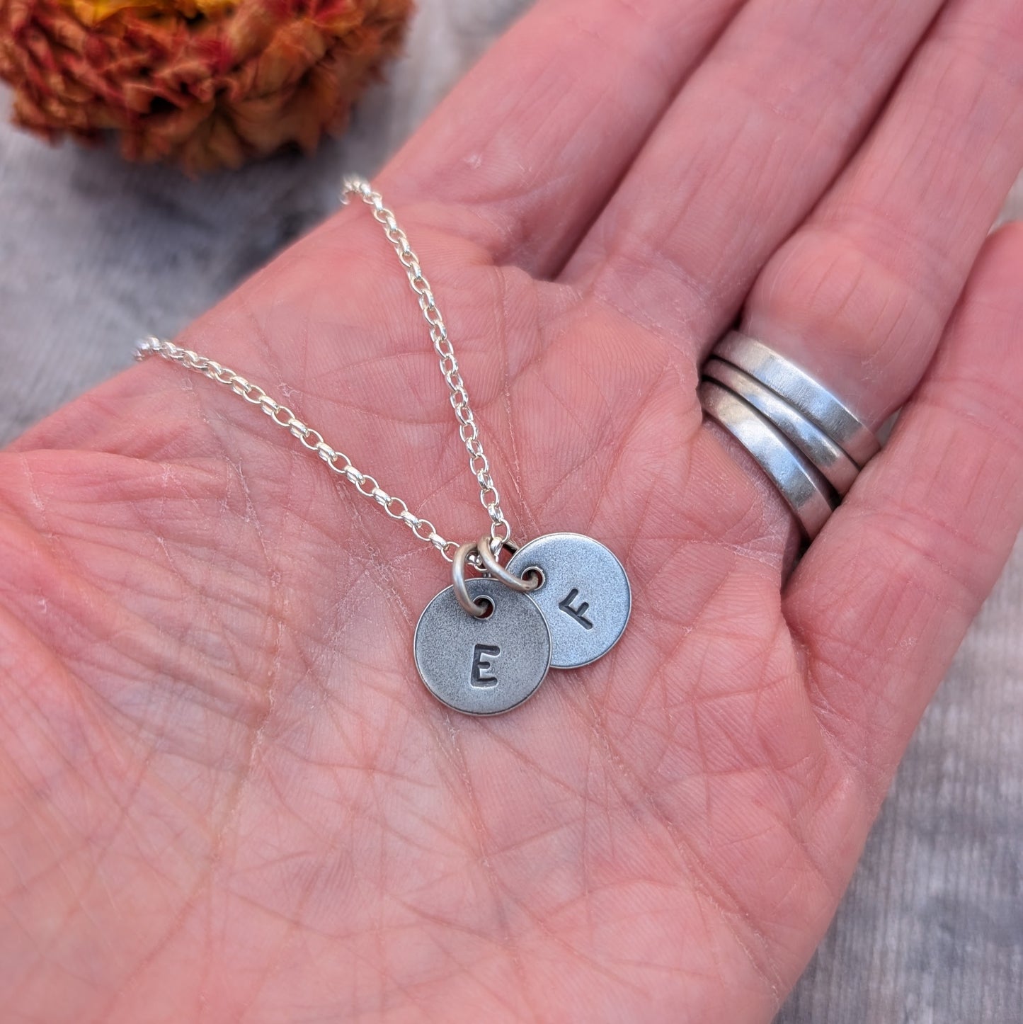 A hand holding a silver initial necklace with two discs stamped with the letters "E" and "F."