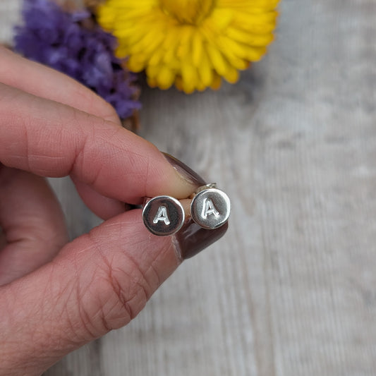 Pair of silver initial stud earrings with the letter “A,” held between fingers with yellow and purple flowers in the background.