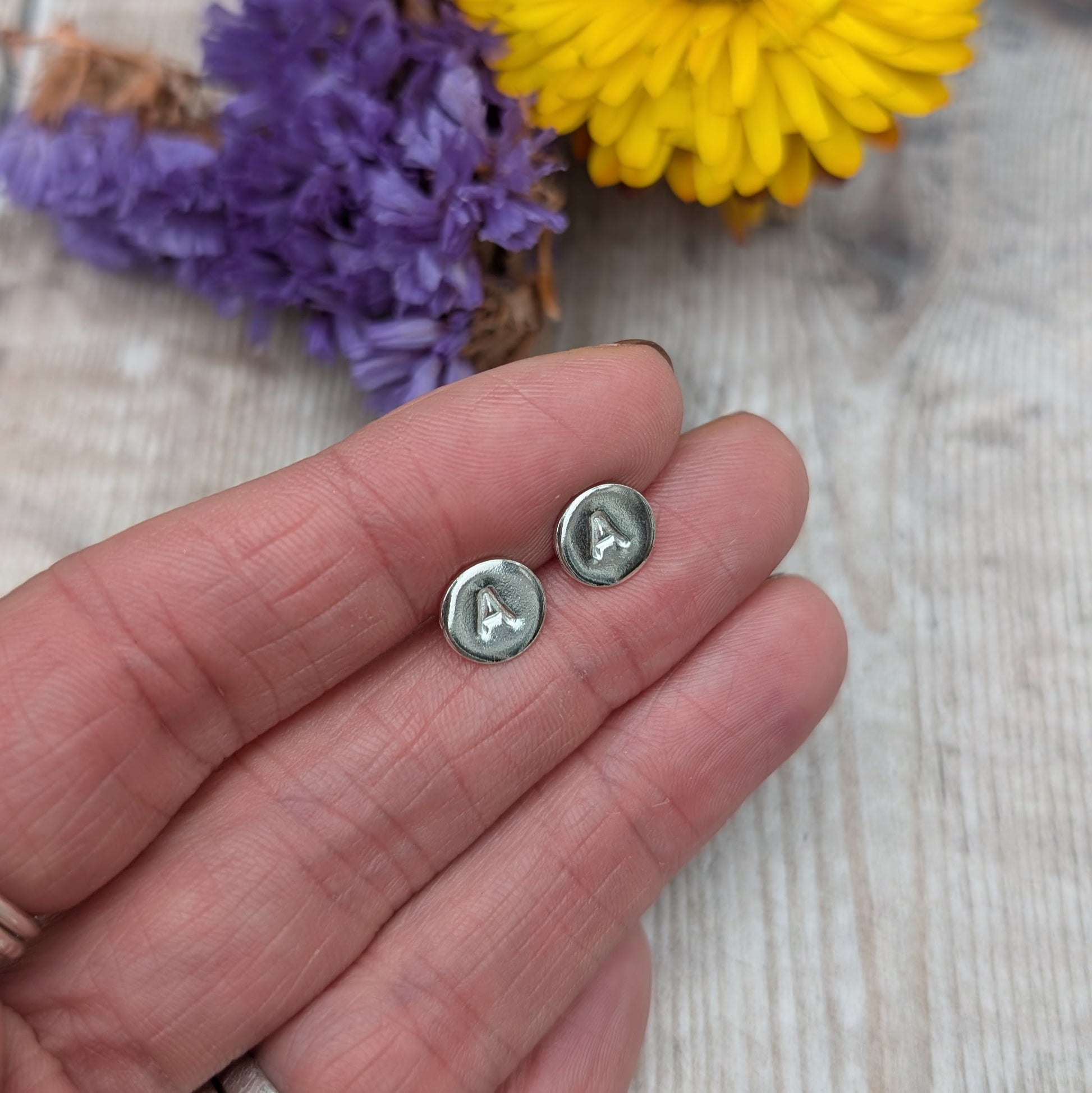 Silver initial stud earrings resting on a hand, showing the stamped letter “A” on each round stud.