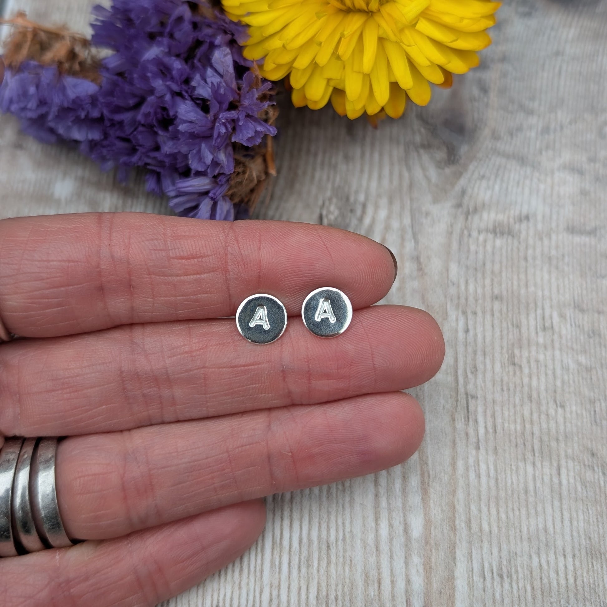 Close-up of silver initial stud earrings on fingers with flowers behind.