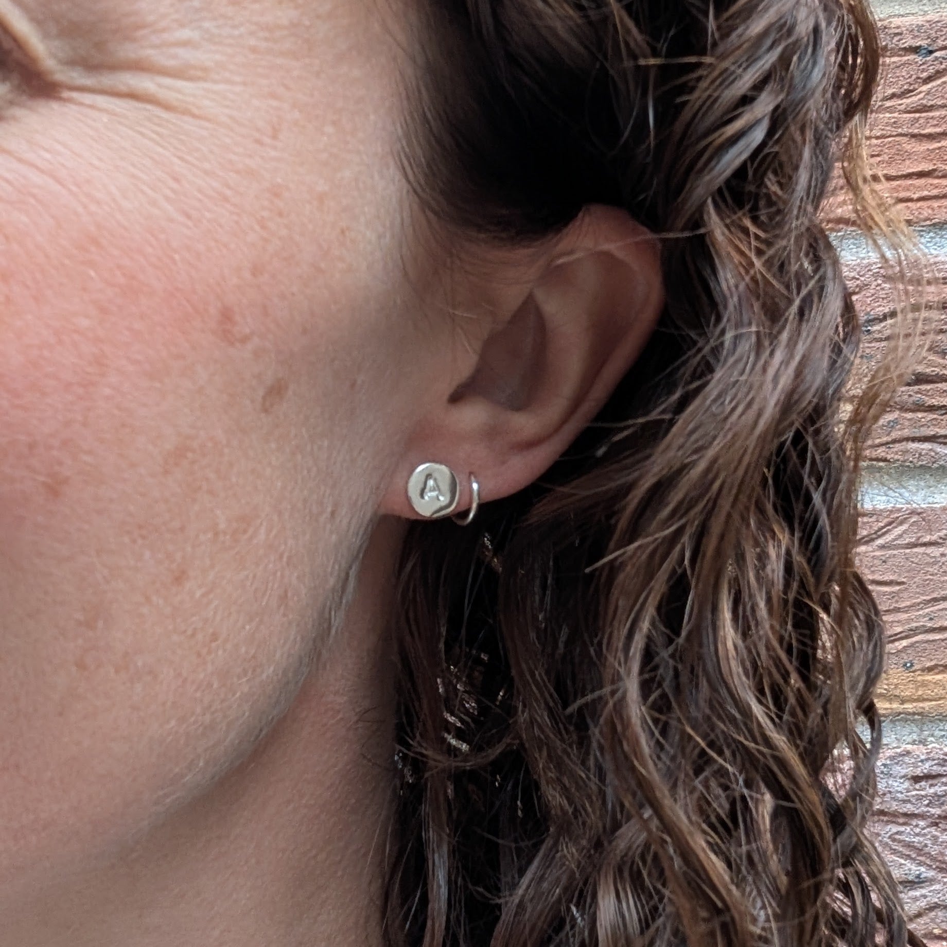 Silver initial stud earring worn on the ear, showing its small circular shape.