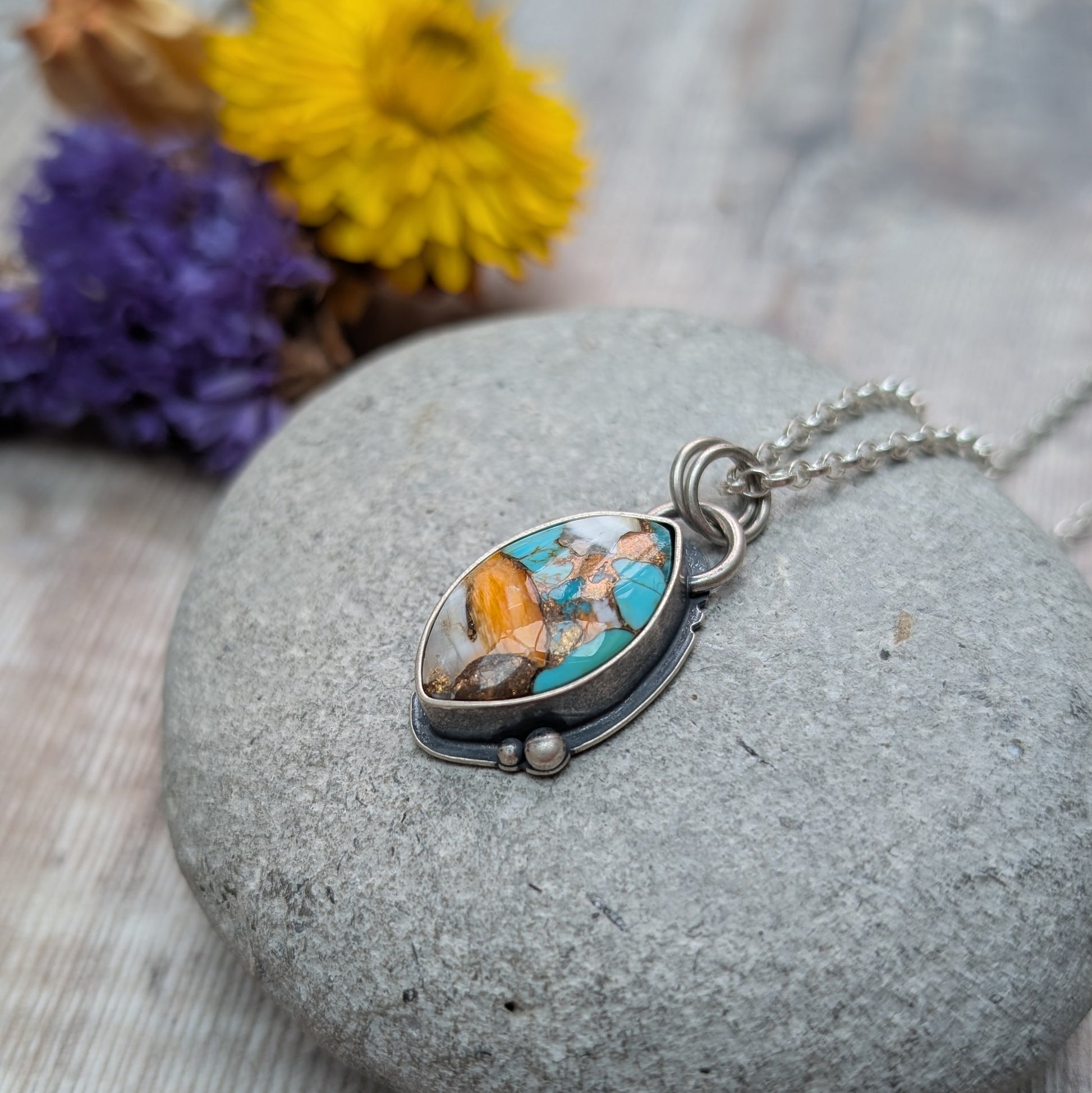 Silver Kingman Turquoise Necklace displayed on a smooth grey stone, with its marquise-shaped multicolour stone setting in oxidised silver, surrounded by blurred yellow and purple flowers.