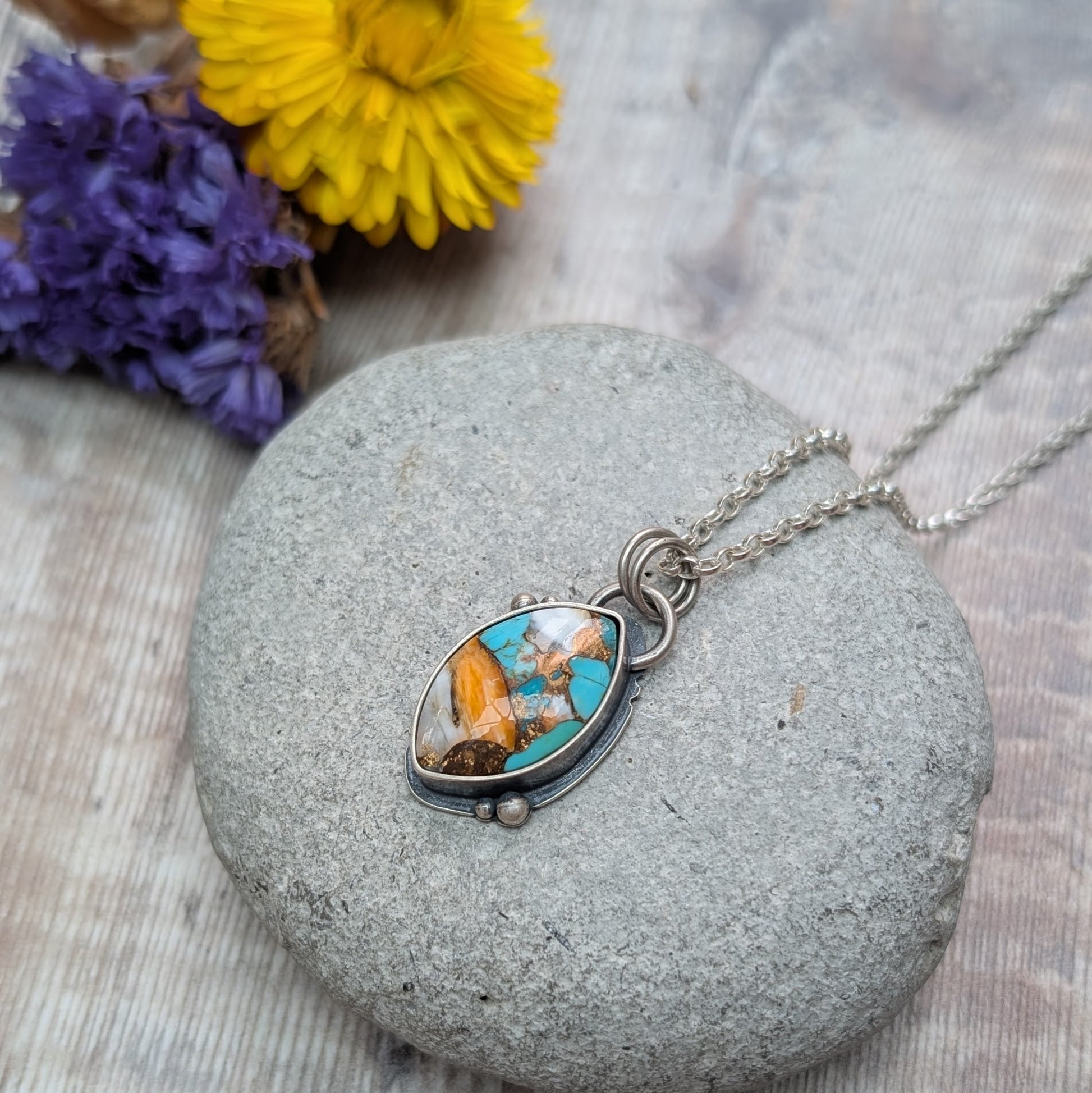 Silver Kingman Turquoise Necklace displayed on a smooth grey stone, with its marquise-shaped multicolour stone setting in oxidised silver, surrounded by blurred yellow and purple flowers.