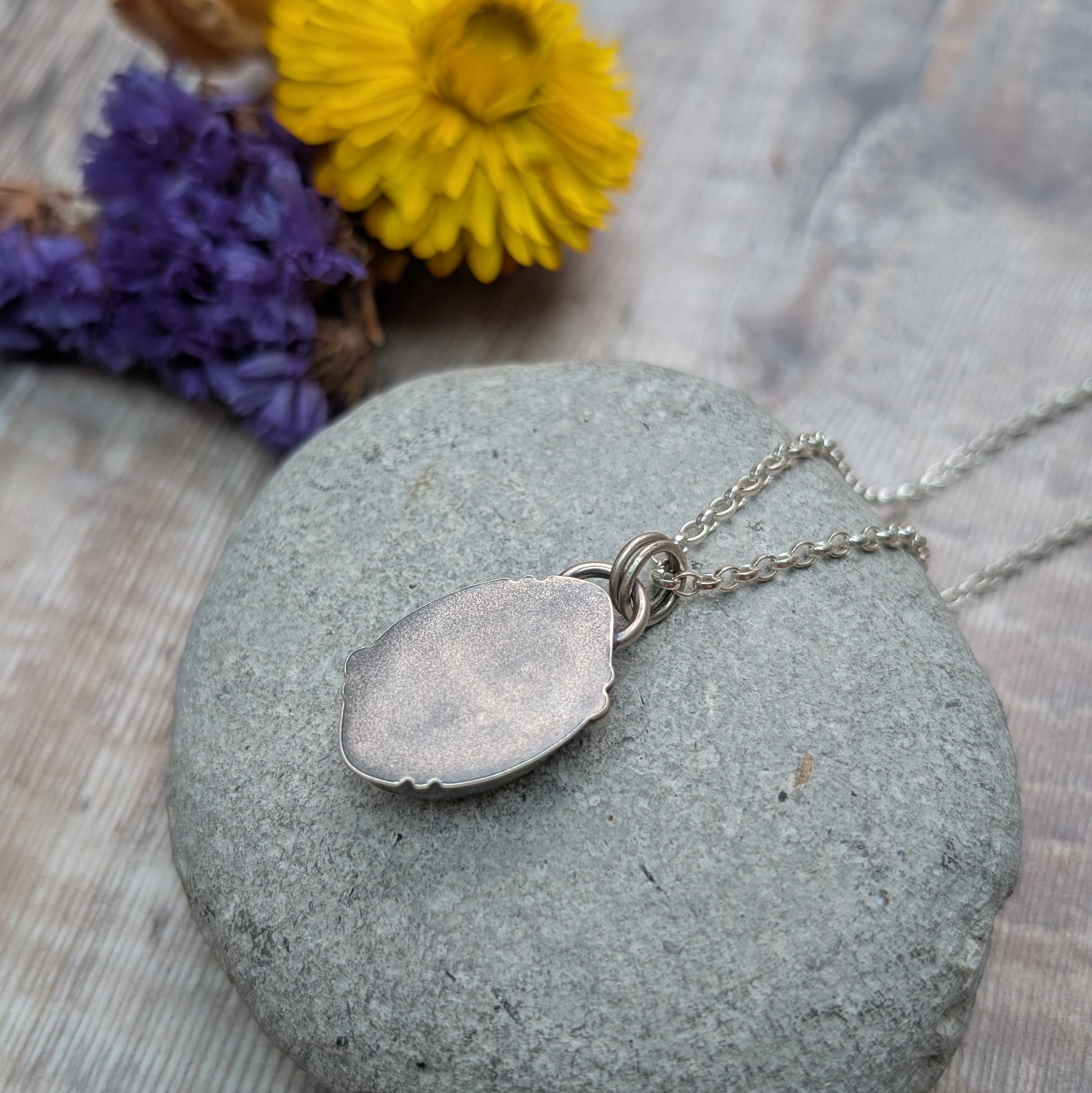 The back of the Silver Kingman Turquoise Necklace pendant, showing the smooth, brushed silver finish and double loop bail, placed on a grey pebble with blurred flowers in the background.