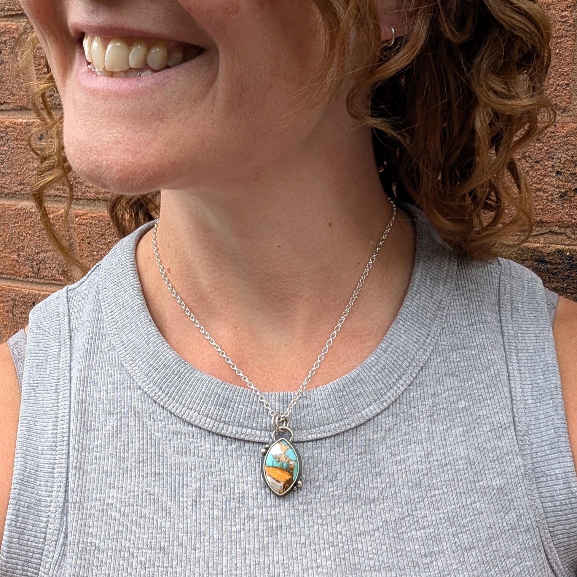 A person wearing the Silver Kingman Turquoise Necklace on a silver chain, with the pendant resting at the collarbone and a subtle smile in view against a brick wall.