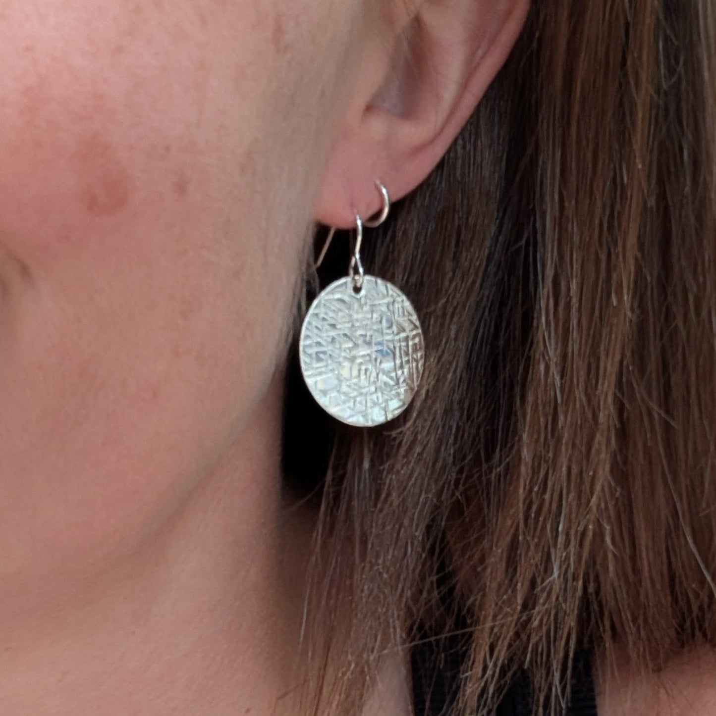 Silver large textured hammered disc earring being worn, showing its round shape and reflective patterned surface.