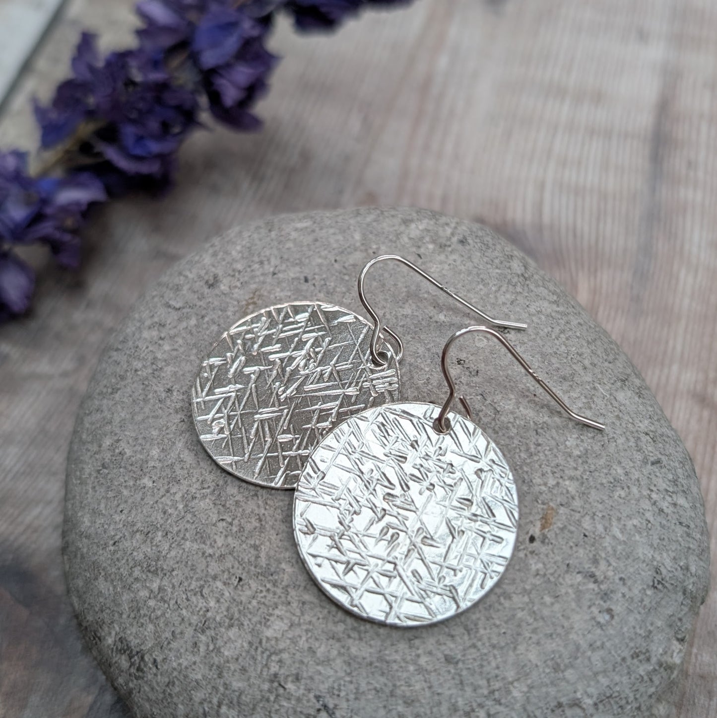 Silver large textured hammered disc earrings displayed on a smooth stone with a blurred floral background.