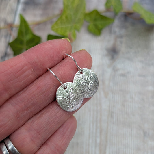 Silver leafy vine oval disc earrings held in a hand, showing the pressed leafy vine pattern and handmade silver ear wires.