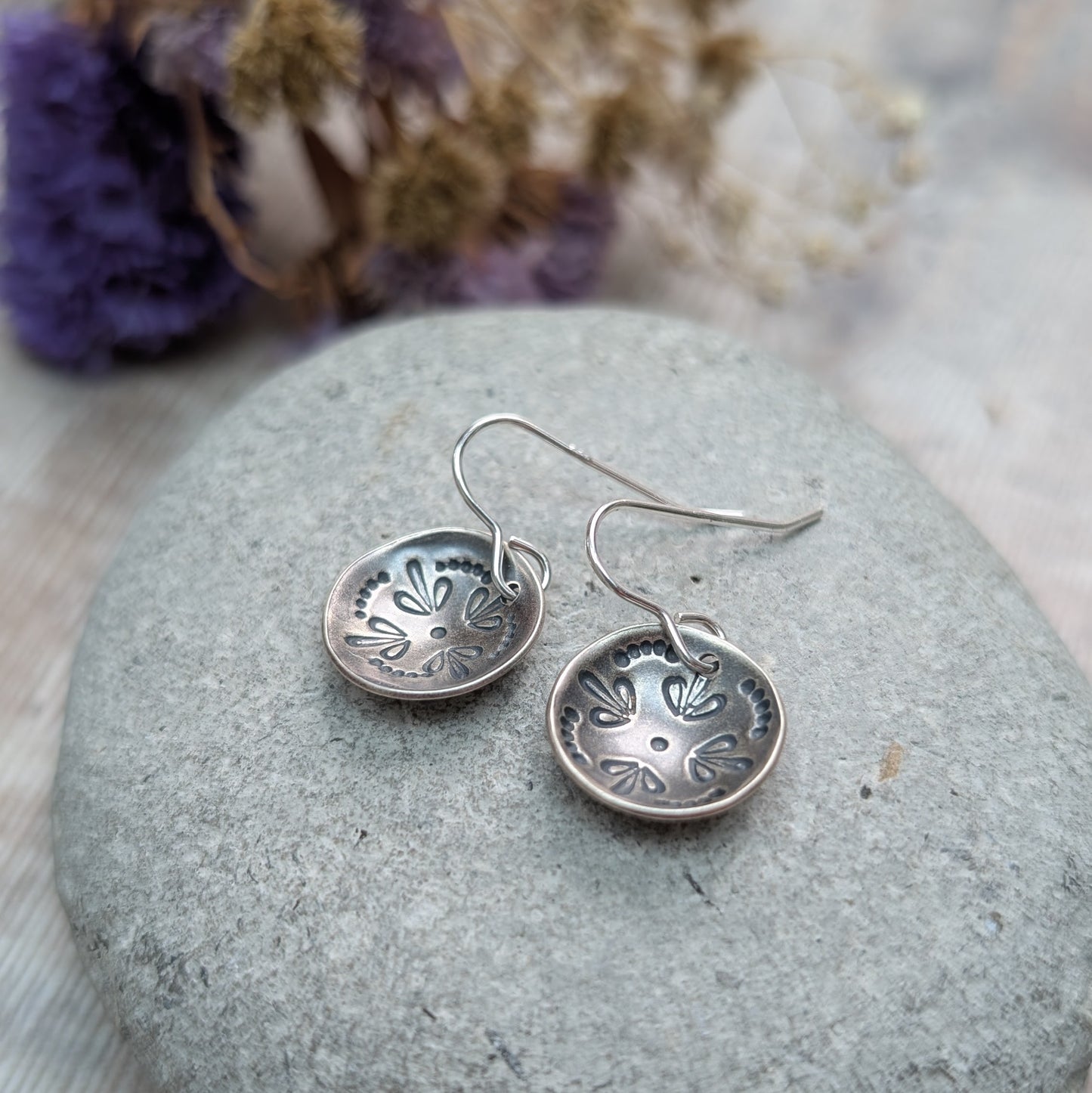 Close-up of a pair of oxidised Silver Mandala Dome Disc Earrings resting on a smooth grey stone, showing the detailed stamped mandala design in the domed surface.