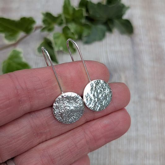 Silver organic disc long earrings held in a hand, showing the textured round silver discs and long curved ear wires.