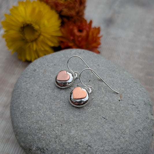 Silver pebble earrings with copper hearts shown close up on a stone, highlighting the polished silver and soft copper detail.