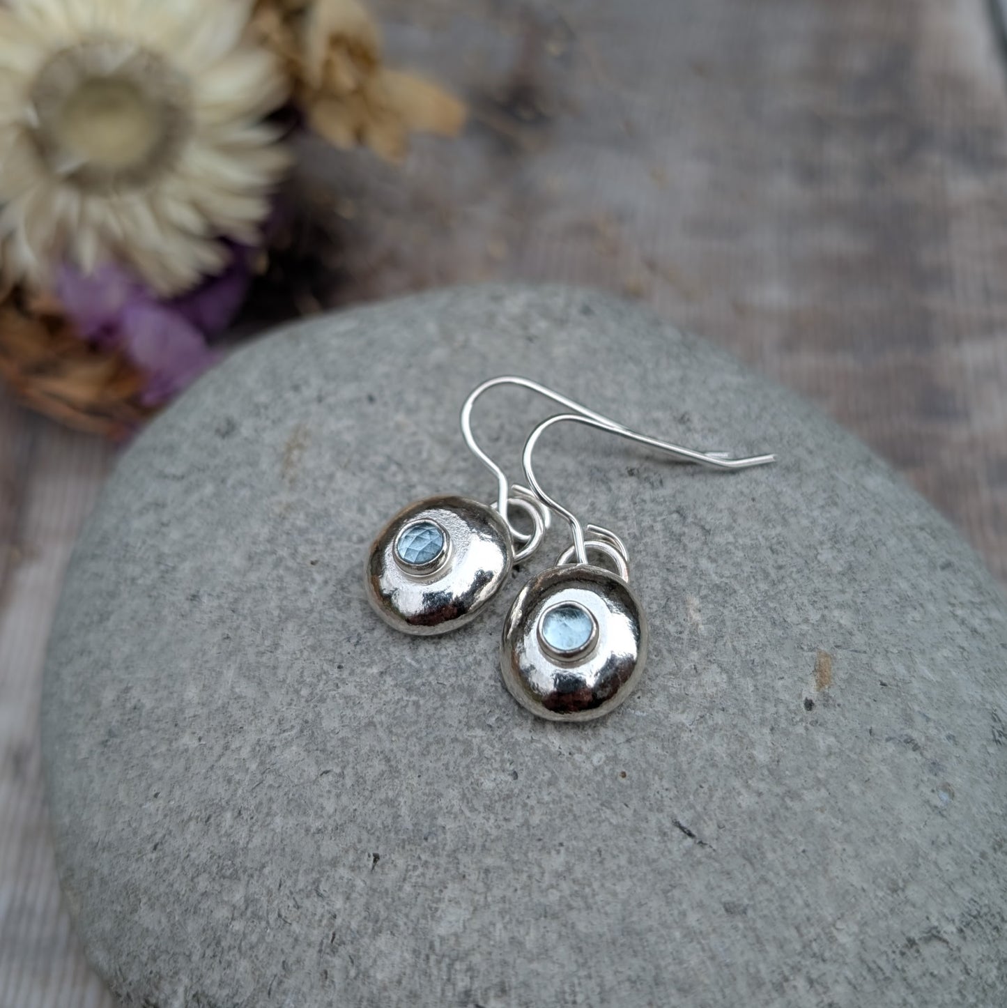 A pair of handmade silver pebble earrings with aquamarine gemstones resting on a smooth stone, highlighting their organic shape and reflective silver finish.