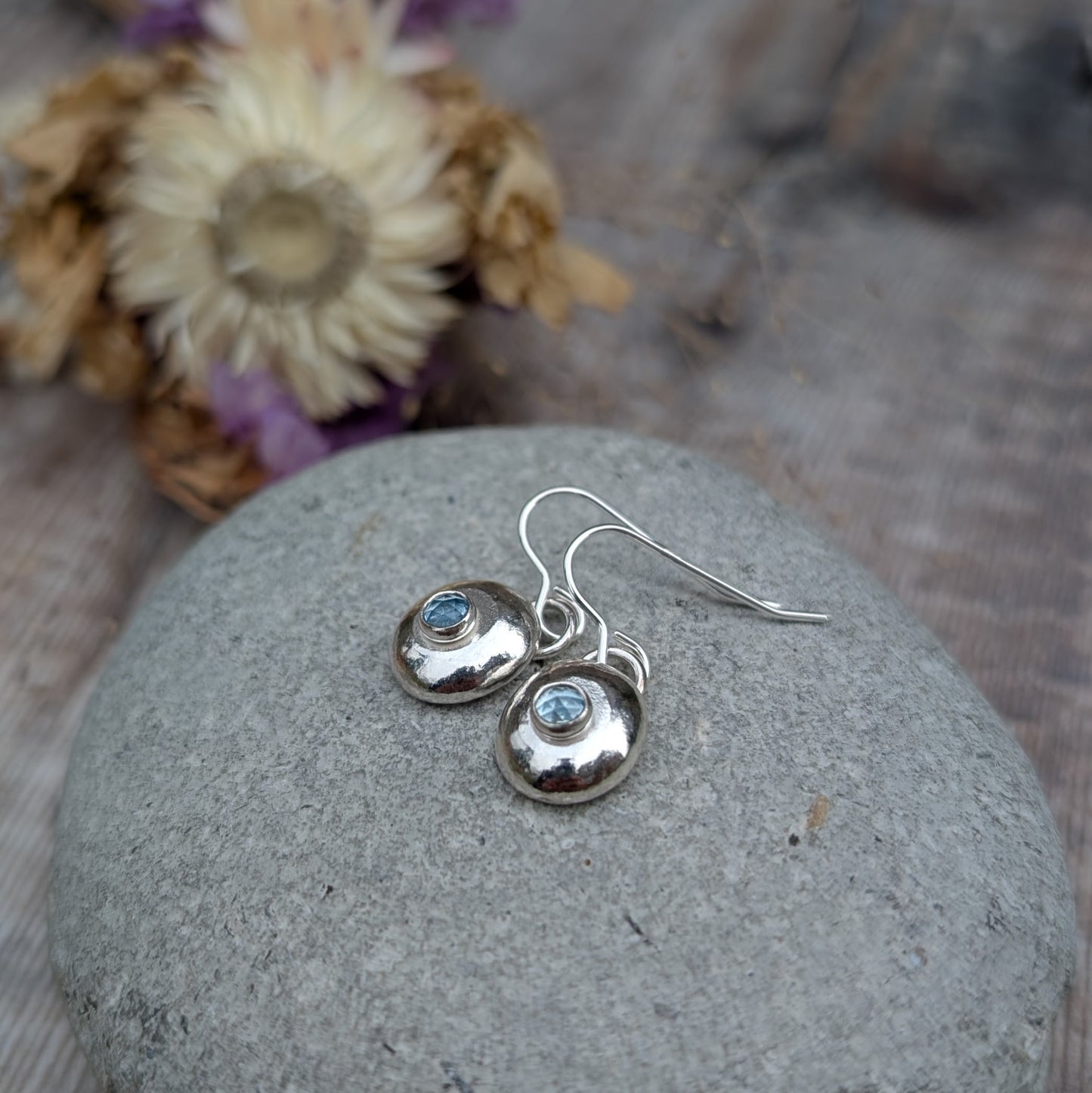 A pair of handmade silver pebble earrings with aquamarine gemstones resting on a smooth stone, highlighting their organic shape and reflective silver finish.