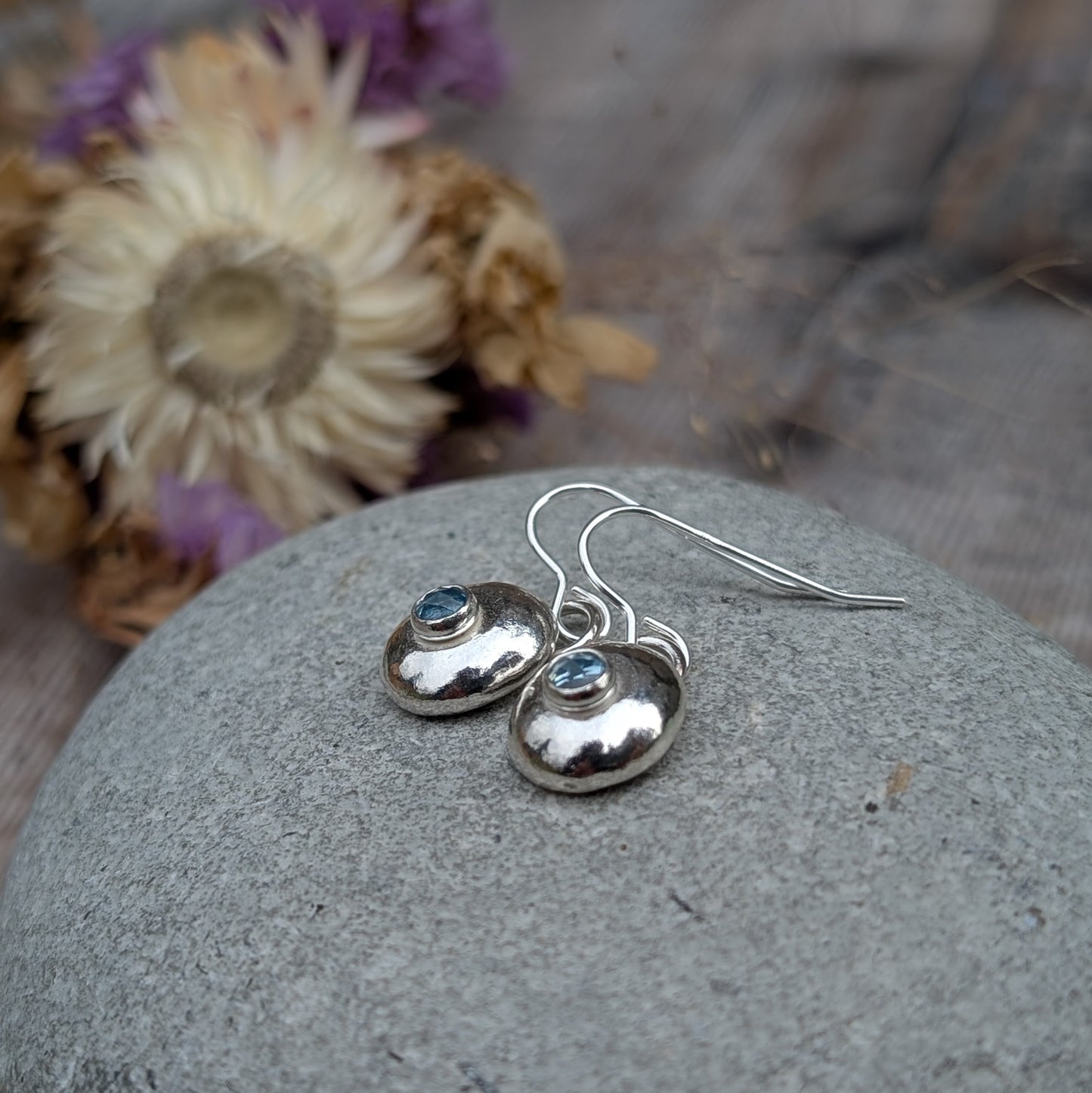 A pair of handmade silver pebble earrings with aquamarine gemstones resting on a smooth stone, highlighting their organic shape and reflective silver finish.