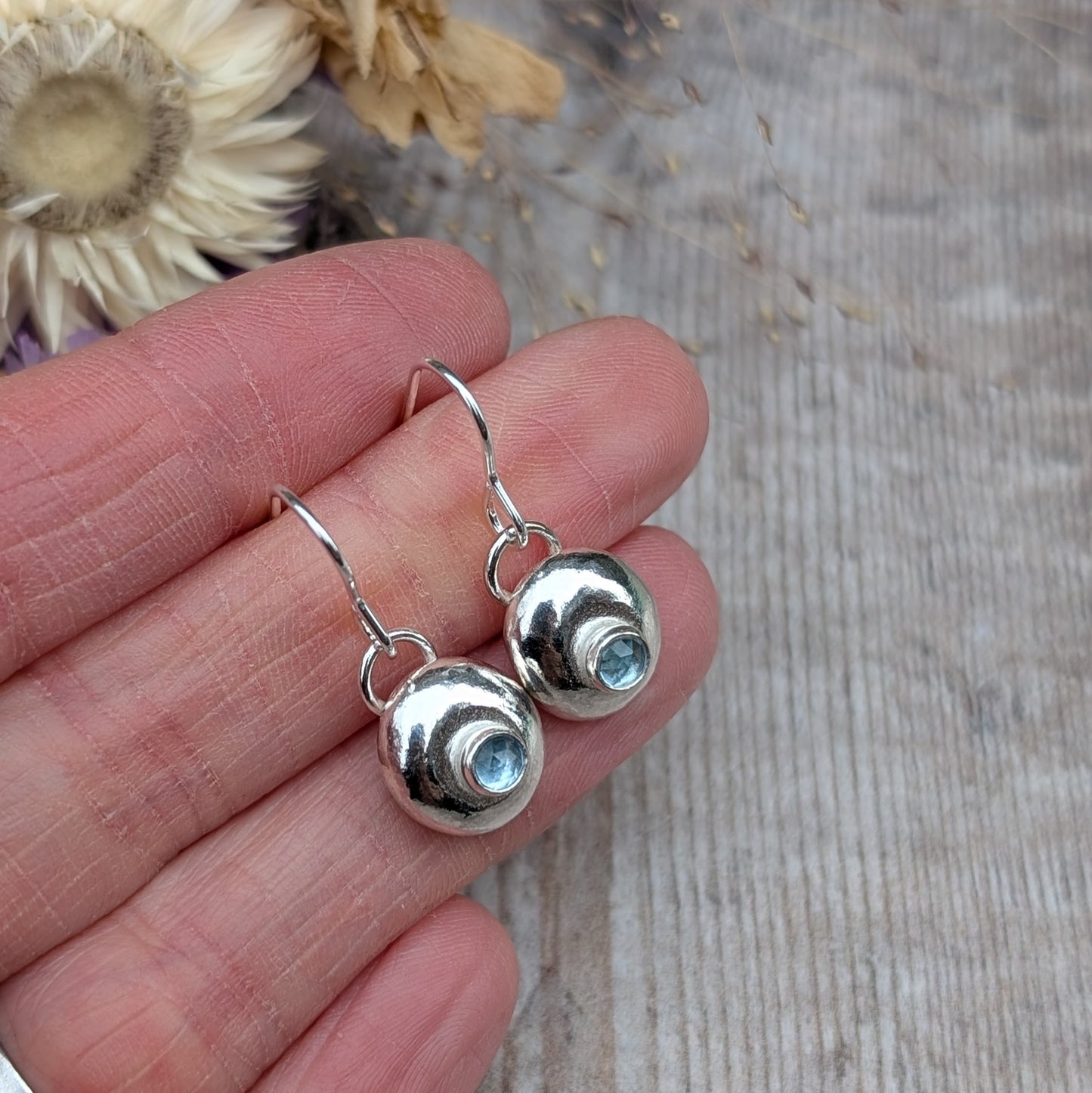 Close-up of a pair of handmade silver pebble earrings with faceted aquamarine stones, held in a person's hand, showcasing their polished surface and organic shape.