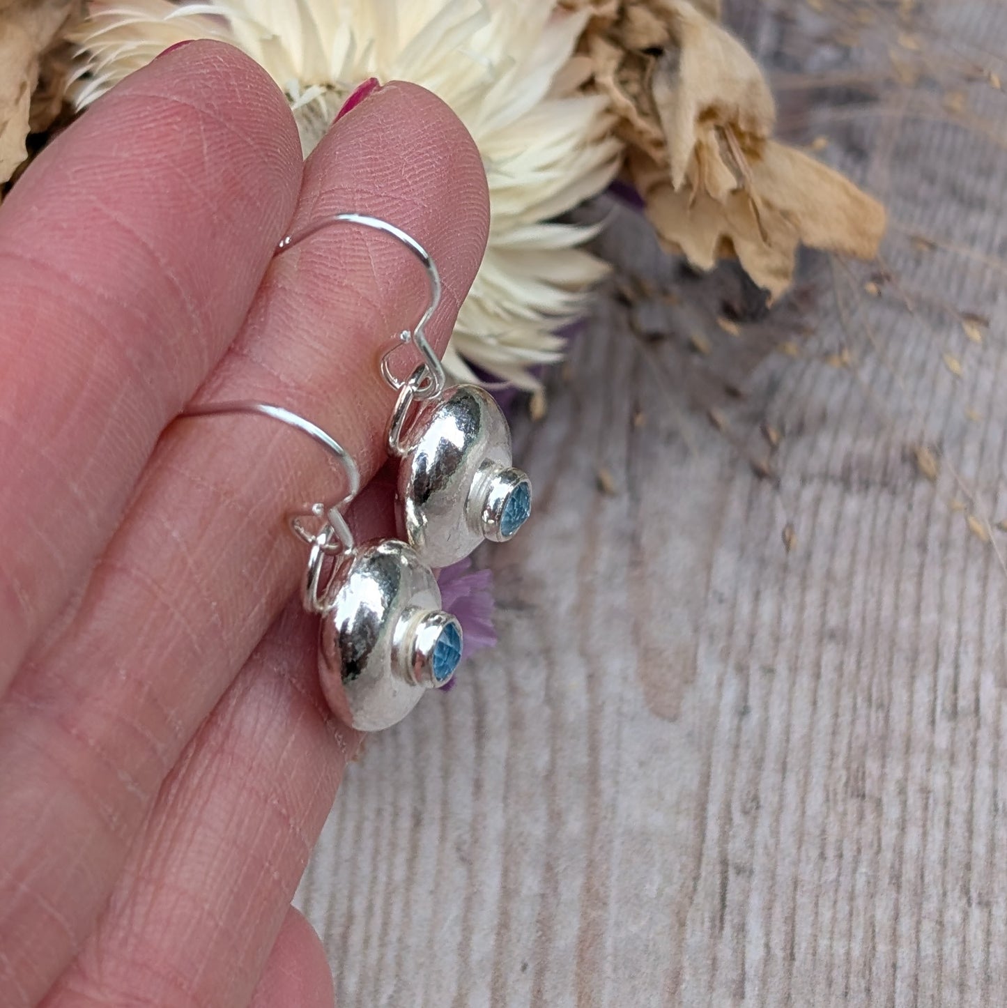 Close-up of a pair of handmade silver pebble earrings with faceted aquamarine stones, held in a person's hand, showcasing their polished surface and organic shape.