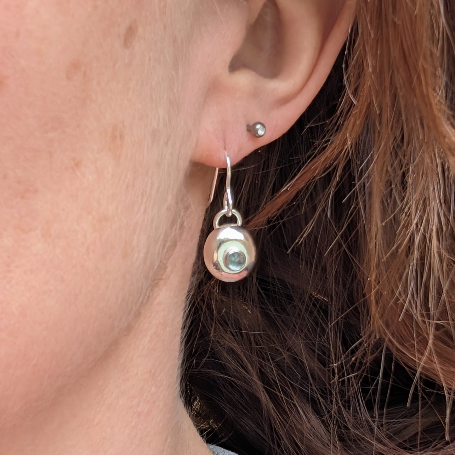 A single handmade silver pebble earring with an aquamarine stone, worn on an ear, showing its size and how it dangles from a silver hook.
