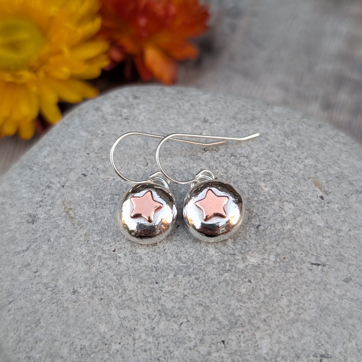 Silver pebble earrings with copper star design, photographed with warm-toned flowers in the background.