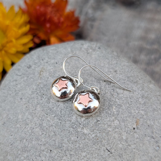 Handmade silver pebble drop earrings with small copper star details, shown resting on a grey stone.