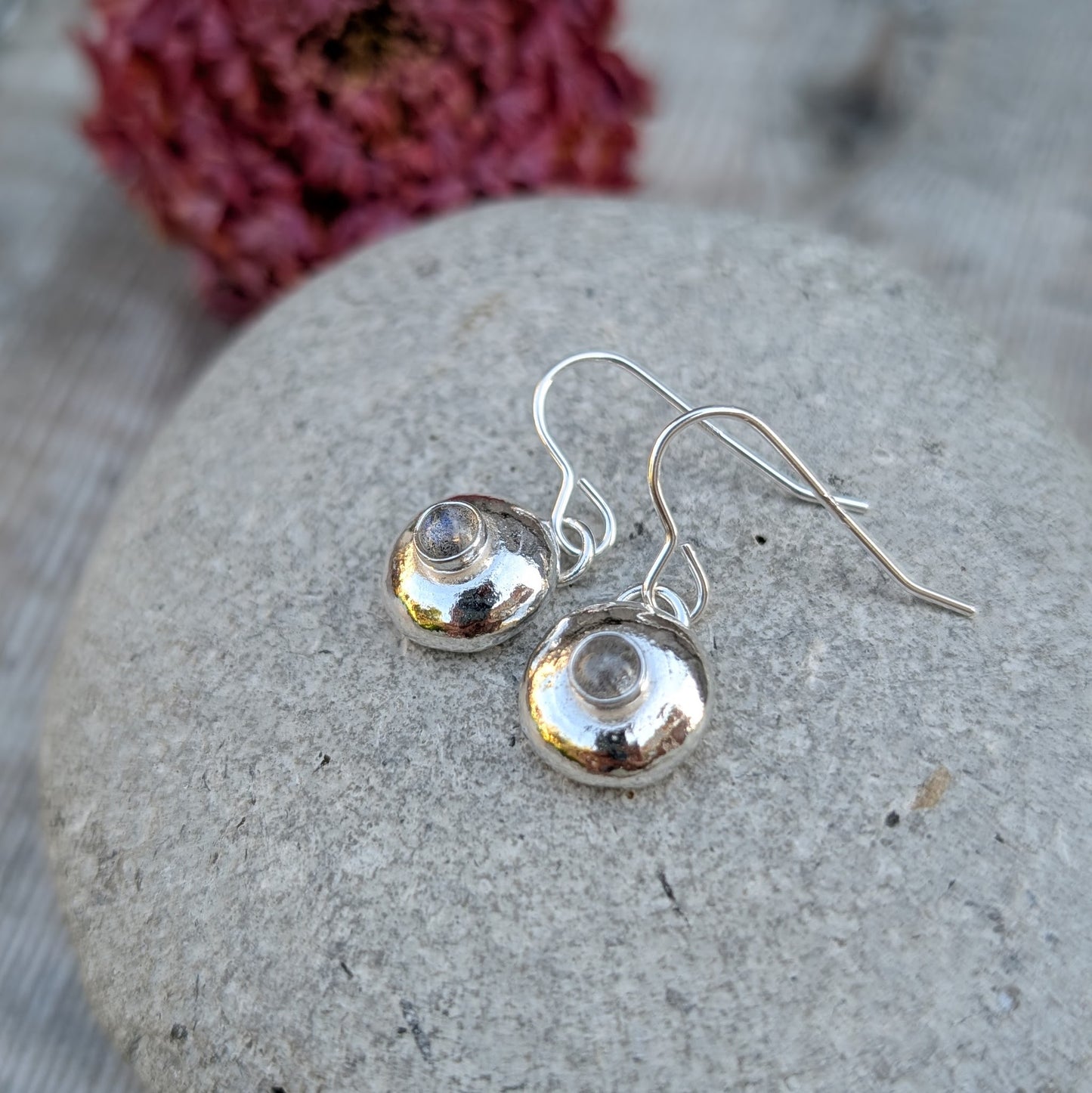 Close-up of silver pebble earrings with Labradorite on a stone, with a soft red flower blurred behind.
