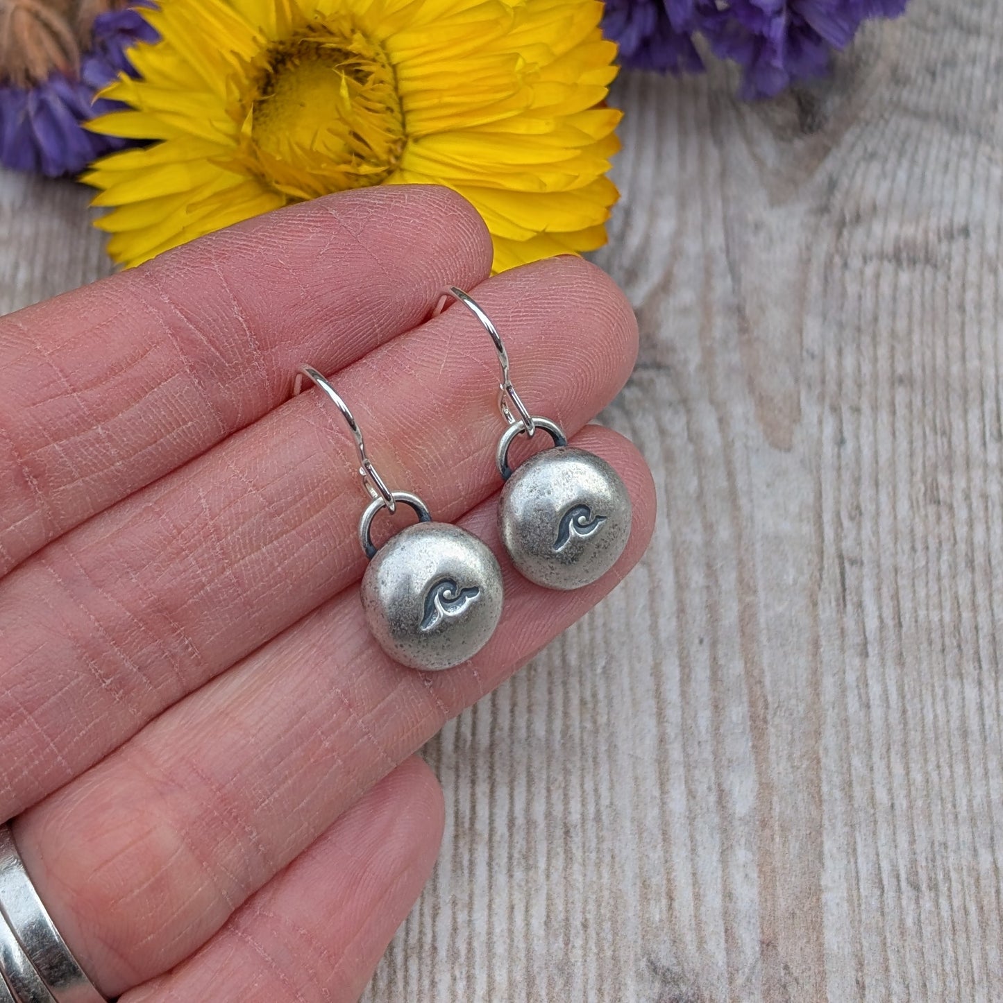 A close-up of a pair of handmade recycled sterling silver pebble earrings with a stamped wave design, held in a hand with a yellow flower in the background.