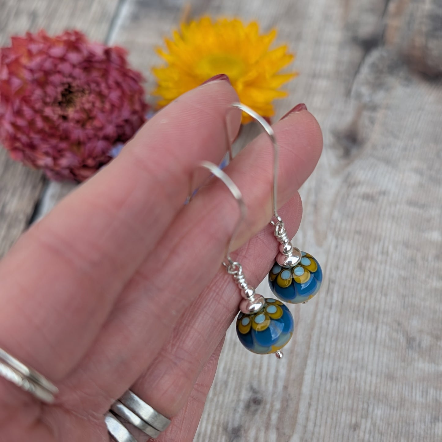 Silver petals lampwork glass bead earrings held up against a wooden background with dried flowers, highlighting the glossy finish and petal-like glass design.