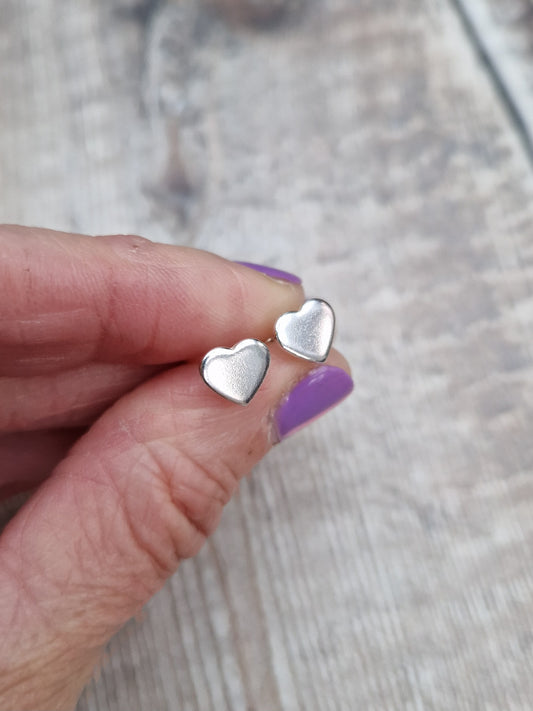 Each earring is a small silver heart shape, flat and smooth, mounted on a silver stud bar.