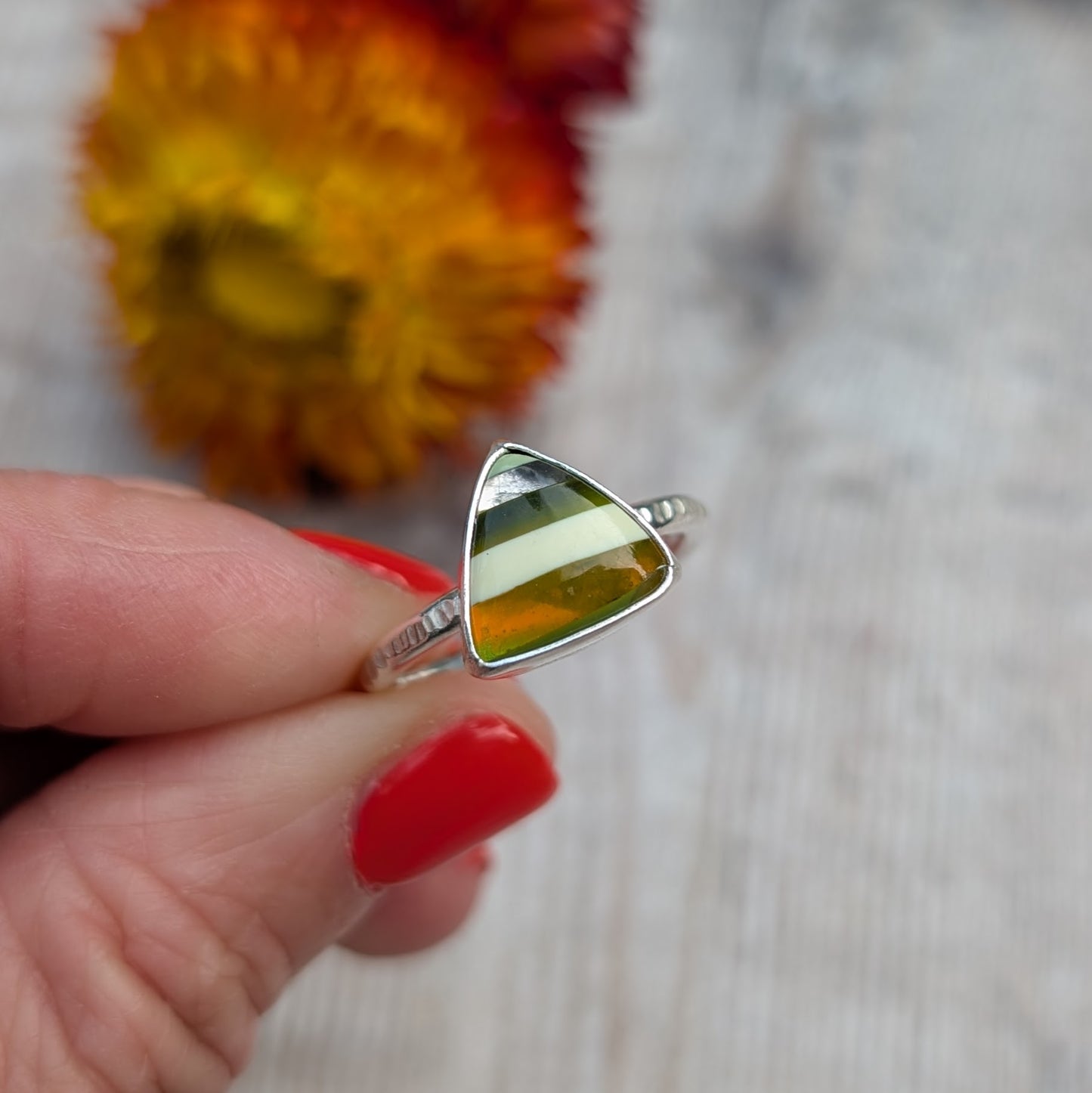 Hand holding a silver ring with a bold, striped Surfite stone, with a blurred flower in the background.