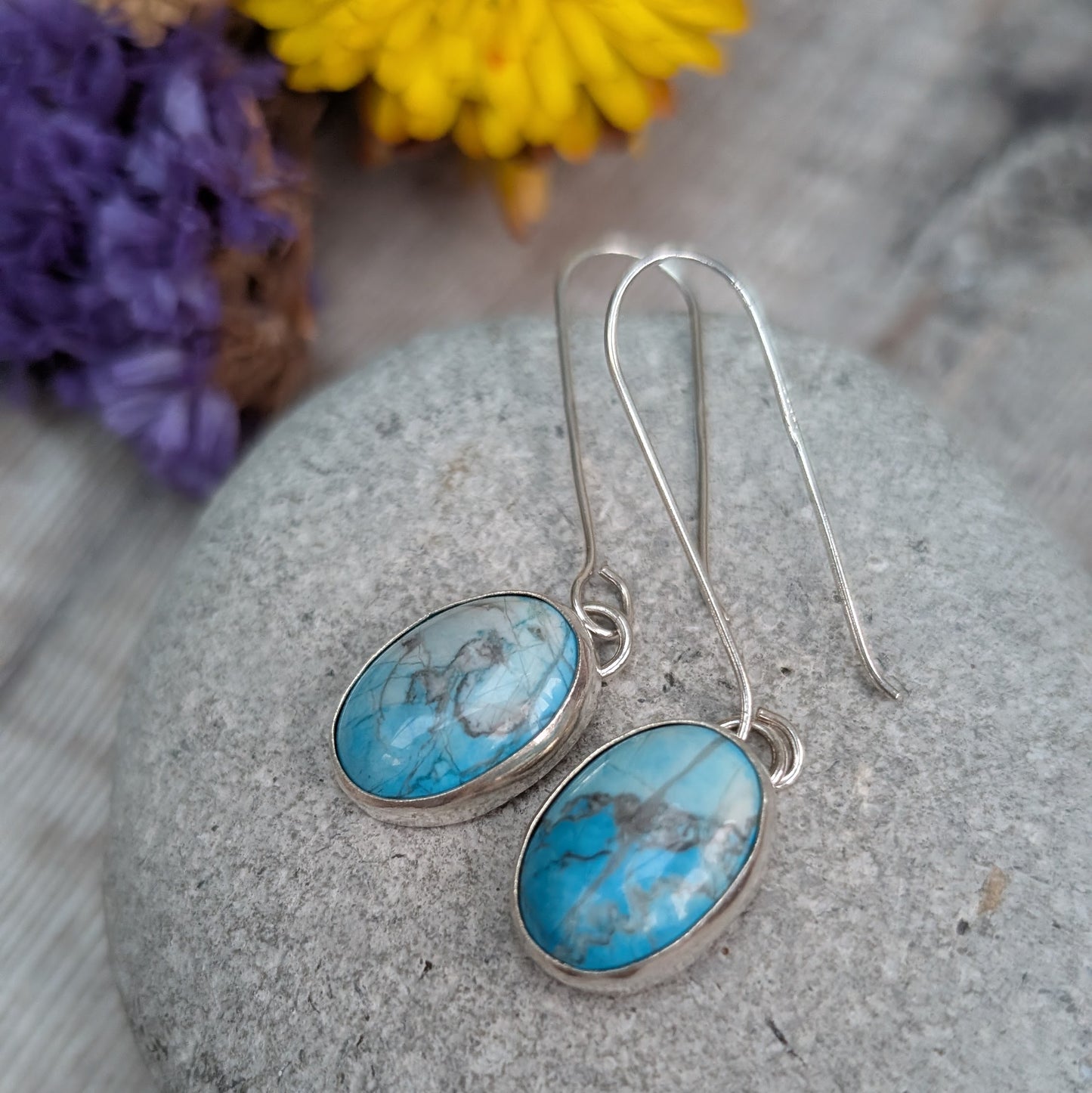 A close-up of the handmade silver Turquoise Howlite earrings on a grey pebble, with the blue stones and natural veining clearly visible.