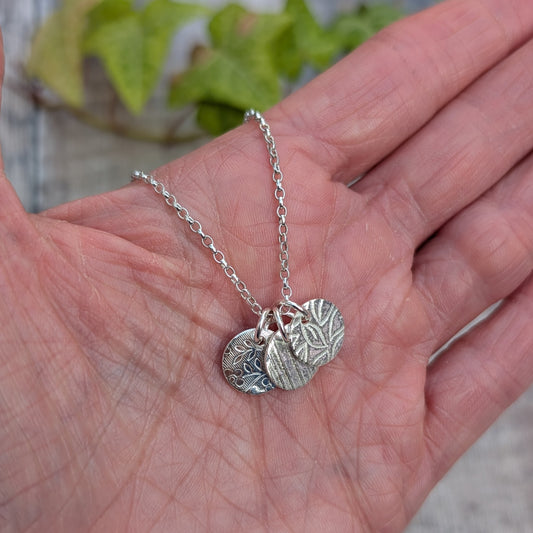Silver wildflower trio of discs necklace handmade from recycled Sterling Silver, shown resting in a hand with three different patterned silver discs.