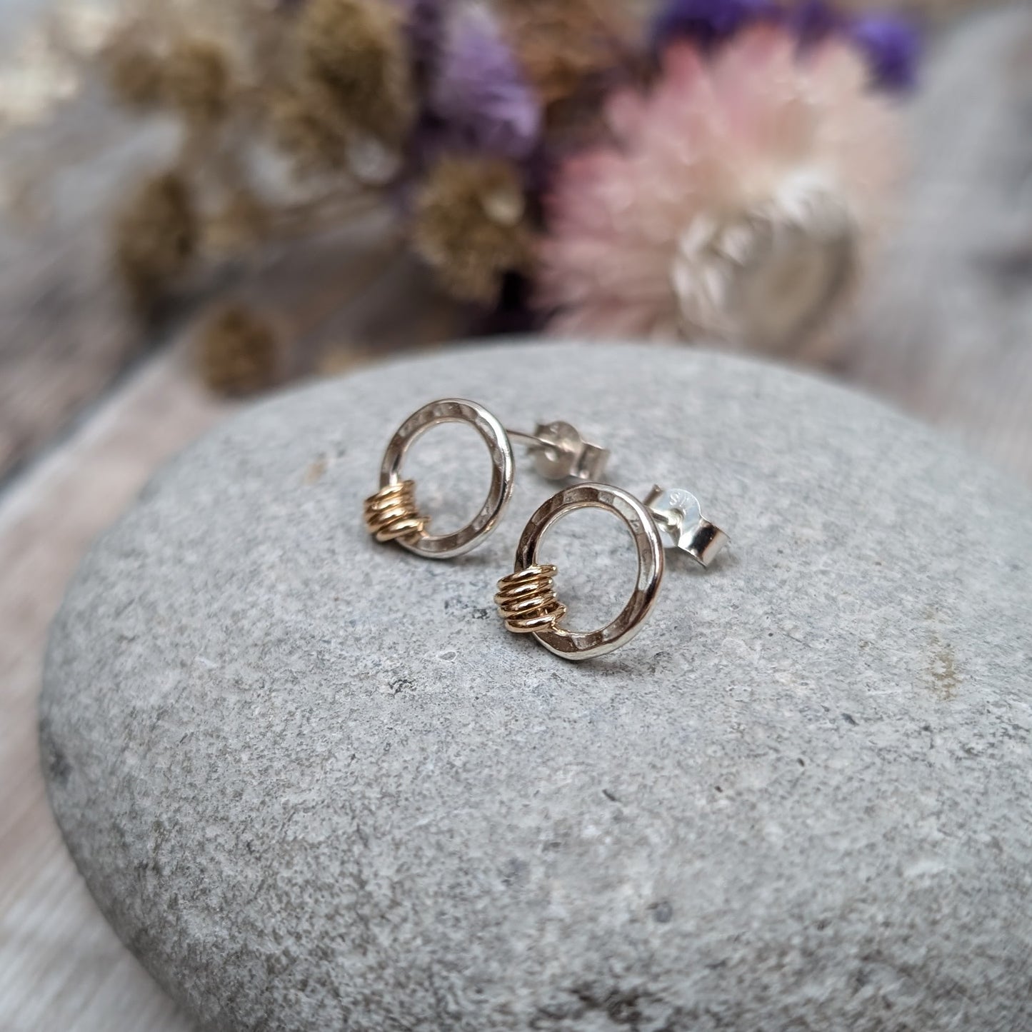 Silver circle stud earring with gold loops shown resting on a grey stone with dried flowers in the background.
