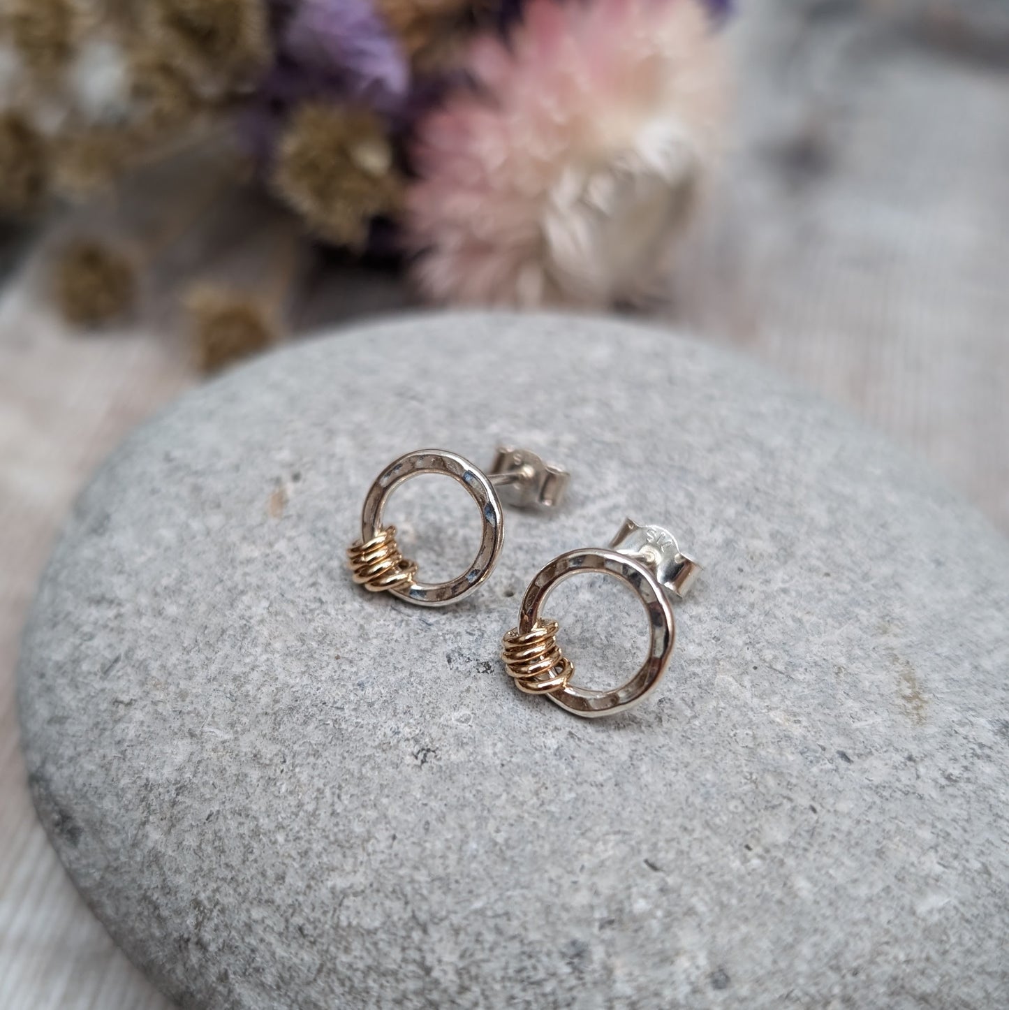 Silver circle stud earrings with gold loops shown displayed on a grey stone with dried flowers in the background.