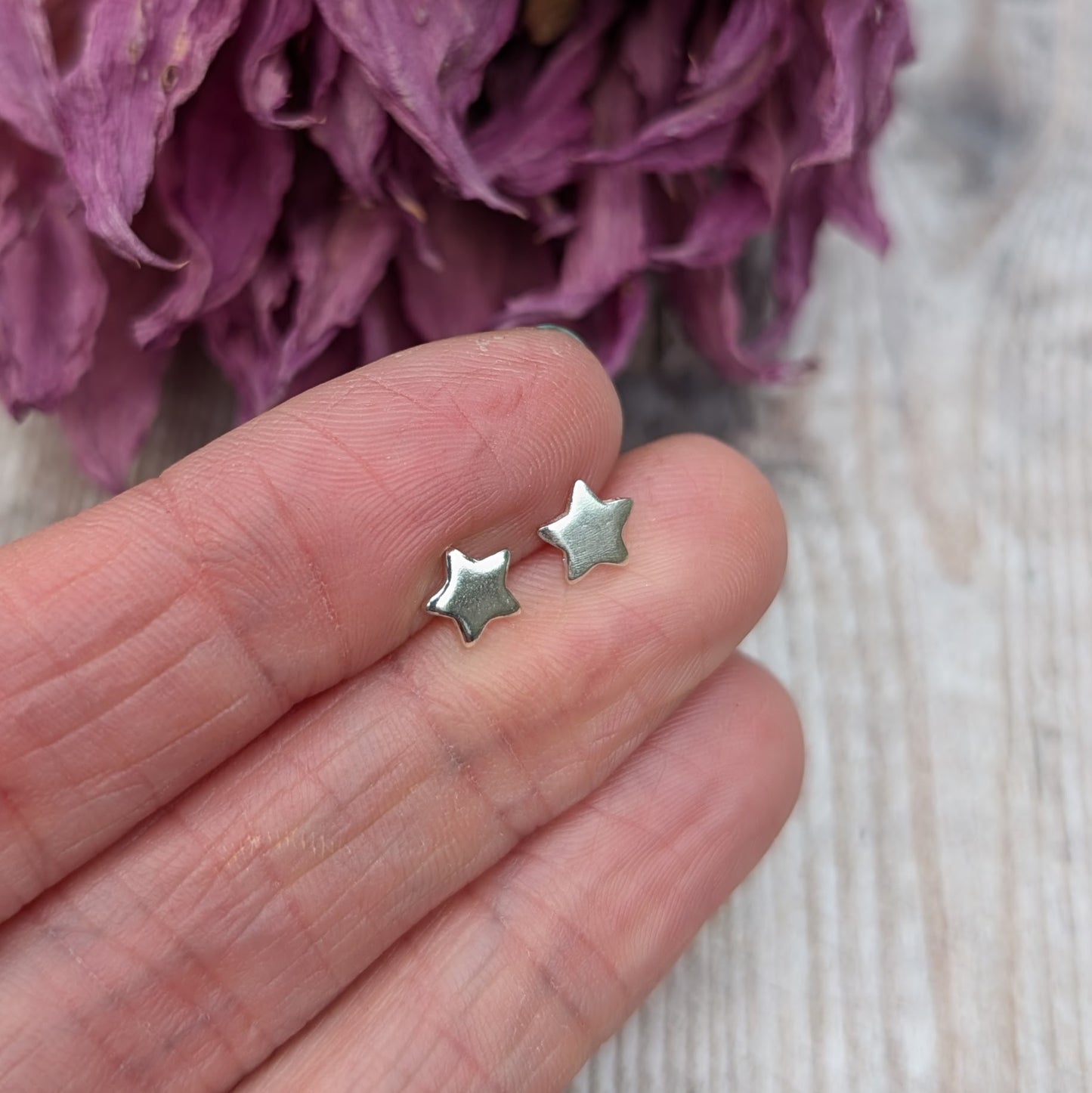 A close-up of a hand holding a pair of small silver star stud earrings, emphasizing their polished finish and minimalist look, with colorful flowers in the background.