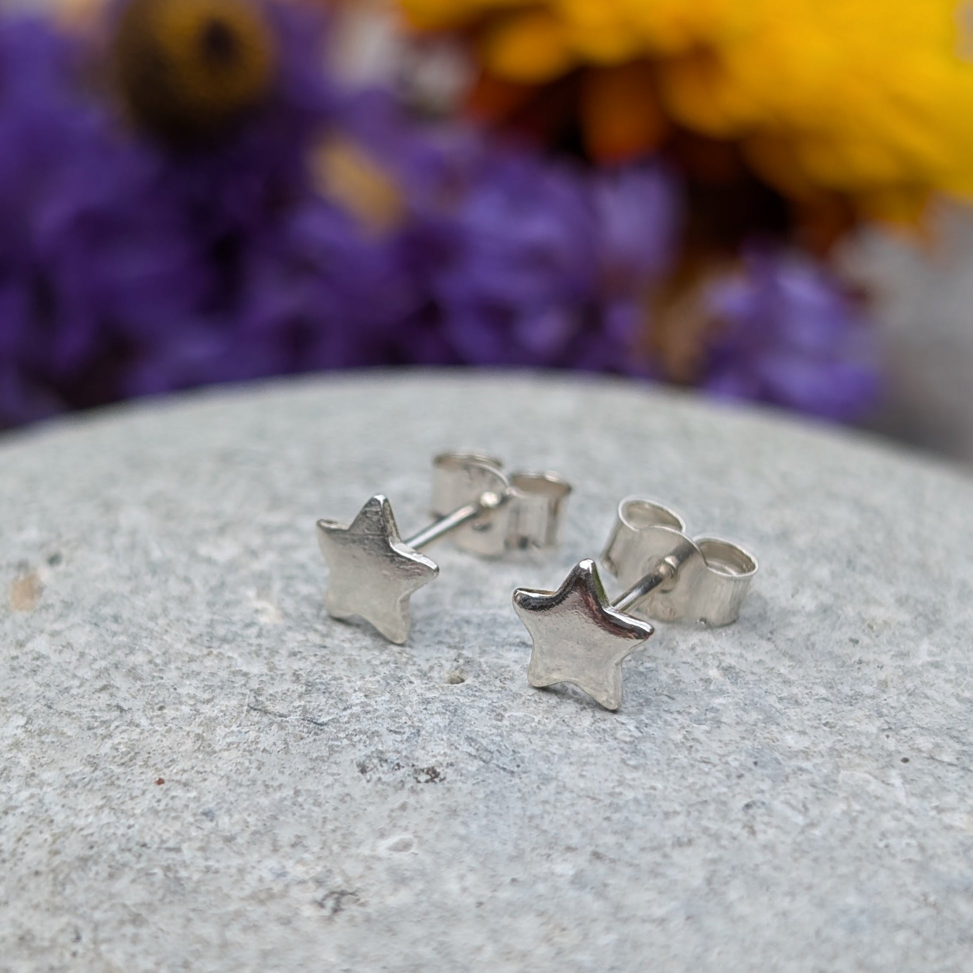 A pair of small silver star stud earrings displayed on a smooth stone surface, with a vibrant floral background in purple and yellow tones.