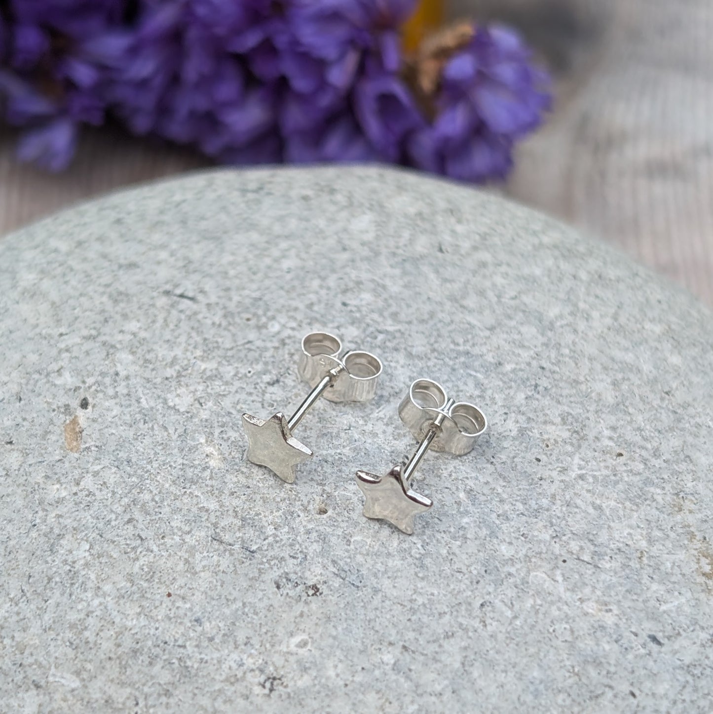A pair of small silver star stud earrings displayed on a smooth stone surface, with a vibrant floral background in purple and yellow tones.