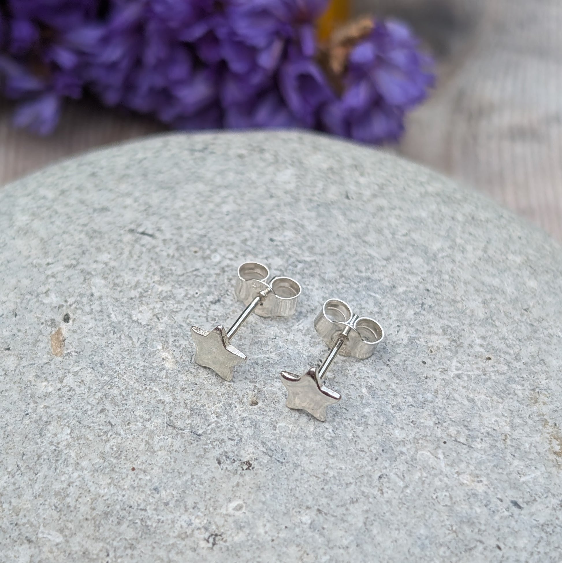 A pair of small silver star stud earrings displayed on a smooth stone surface, with a vibrant floral background in purple and yellow tones.