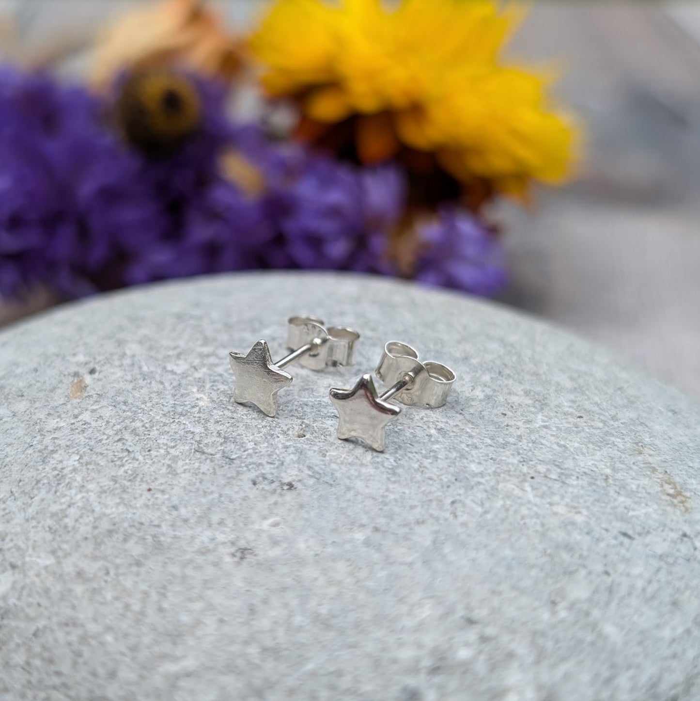 A pair of small silver star stud earrings displayed on a smooth stone surface, with a vibrant floral background in purple and yellow tones.