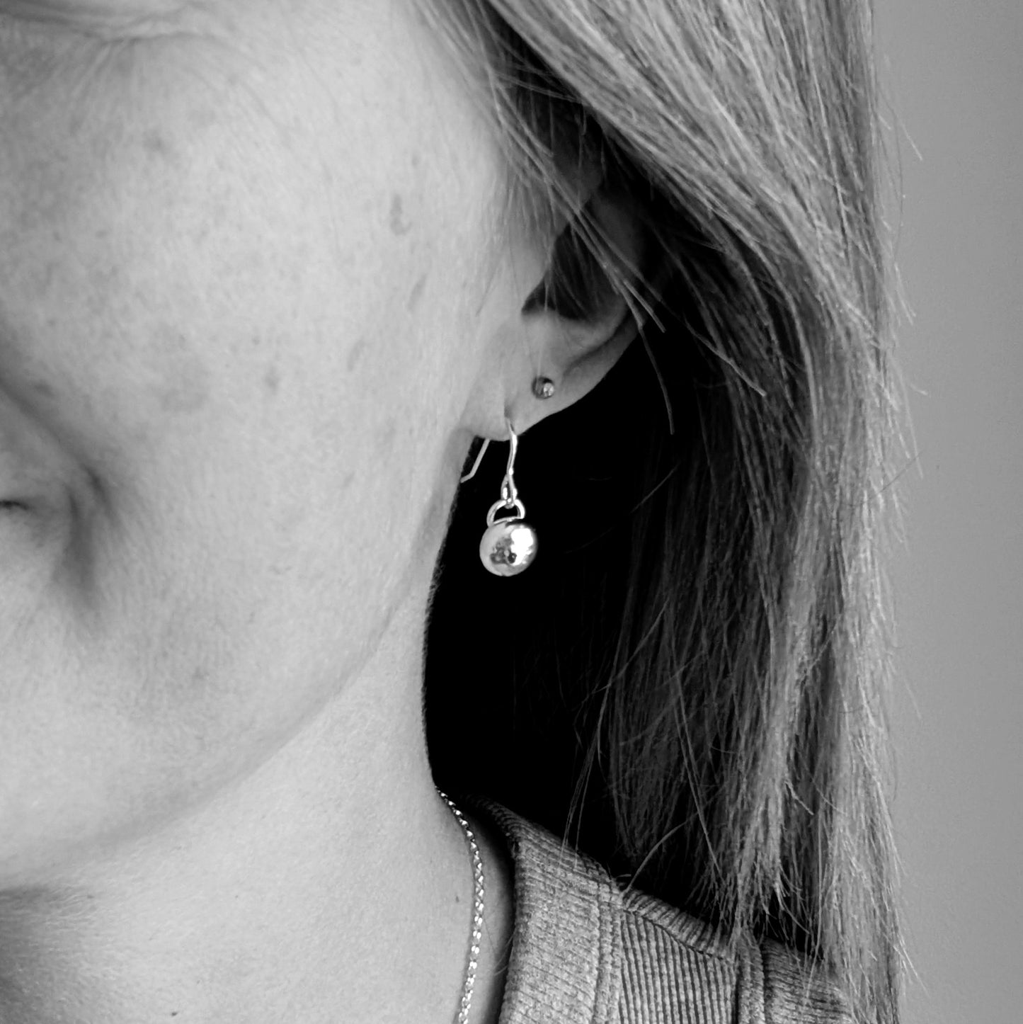 Close-up black and white image of a person wearing small silver dangle earrings with a smooth, pebble-like design. The earrings have a minimalist and modern look, hanging just below the earlobe.