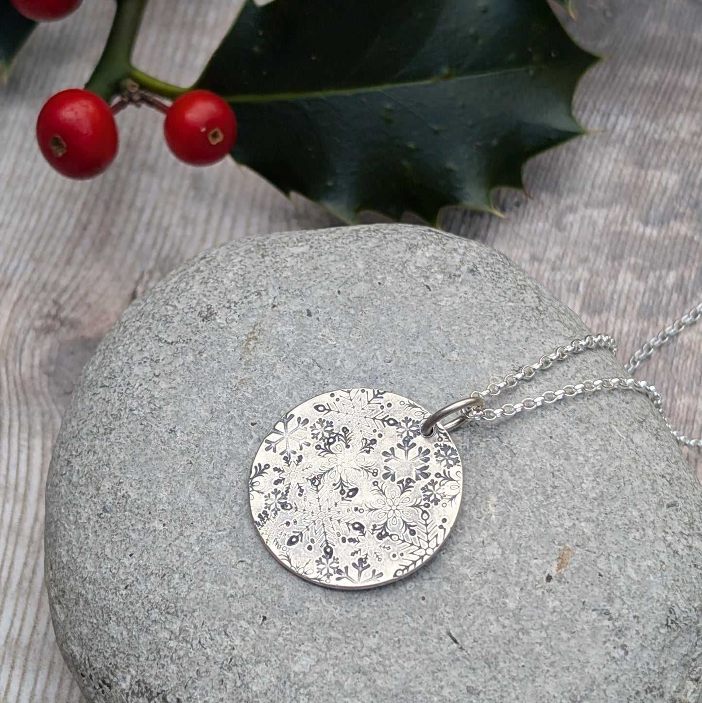 Pendant is a silver disc with intricate stamped snowflakes, some raised and some indented in texture. The snowflakes are randomly stamped and are all different in size and shape. Suspended from an 18 inch silver chain via a small open circle link.